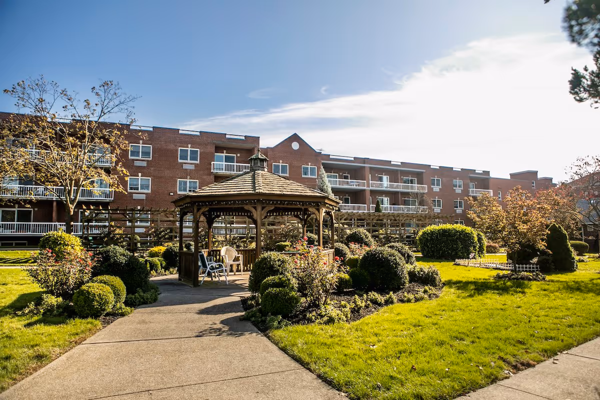 A well-maintained outdoor garden area with a wooden gazebo surrounded by neatly trimmed bushes and flowering plants. In the background, there is a multi-story brick building with balconies and windows. The sky is clear with some clouds, and the sun is shining brightly.