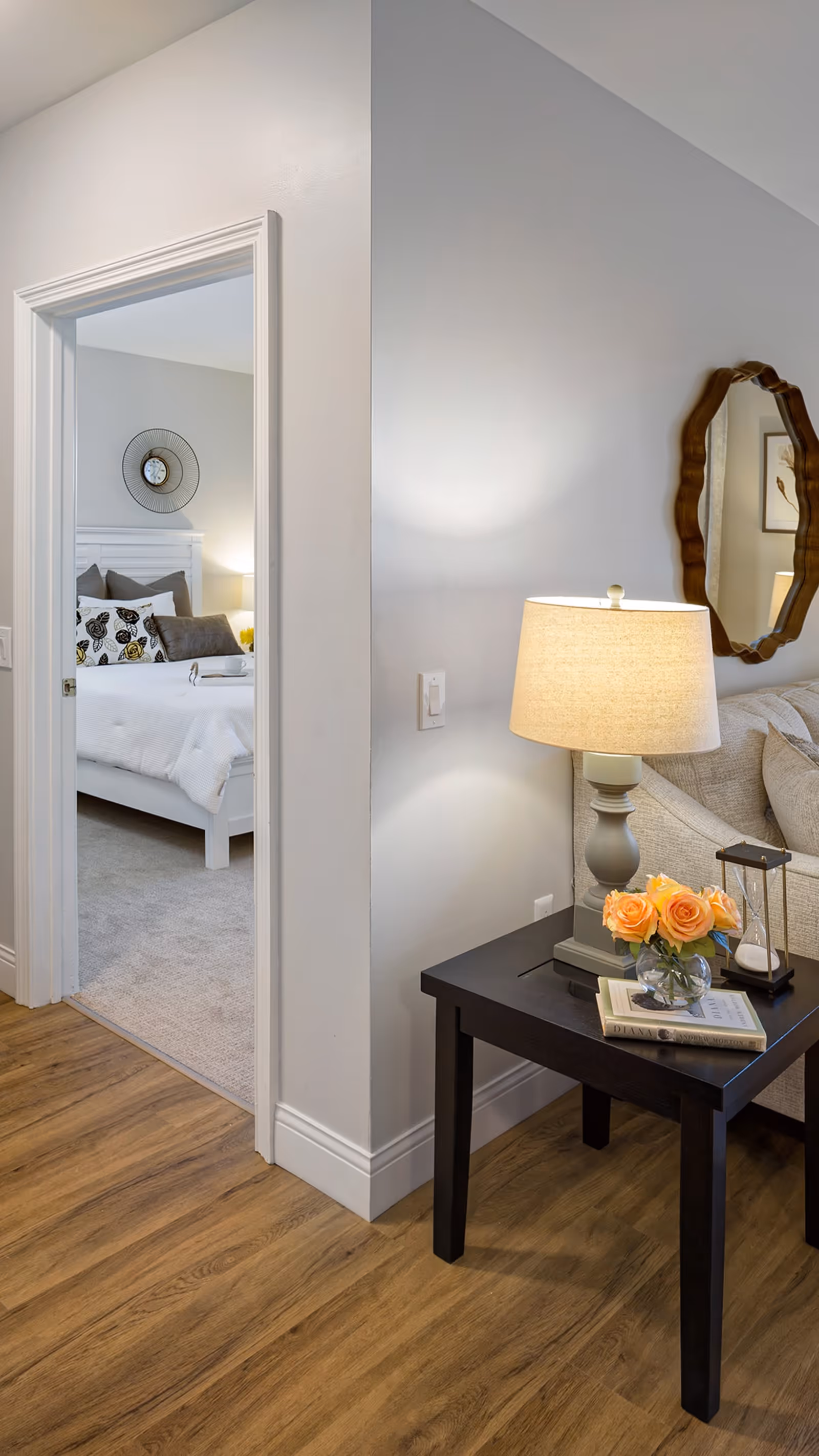 View of a cozy corner in a senior living facility showing a dark wooden side table with a beige lamp, a small vase with orange roses, an hourglass, and two books. To the right is a beige sofa with cushions and a decorative wooden-framed mirror on the wall. To the left, an open doorway reveals a bedroom with a white bed frame, white bedding, decorative pillows, and a wall clock above the headboard. The floor is wood laminate transitioning to carpet in the bedroom.