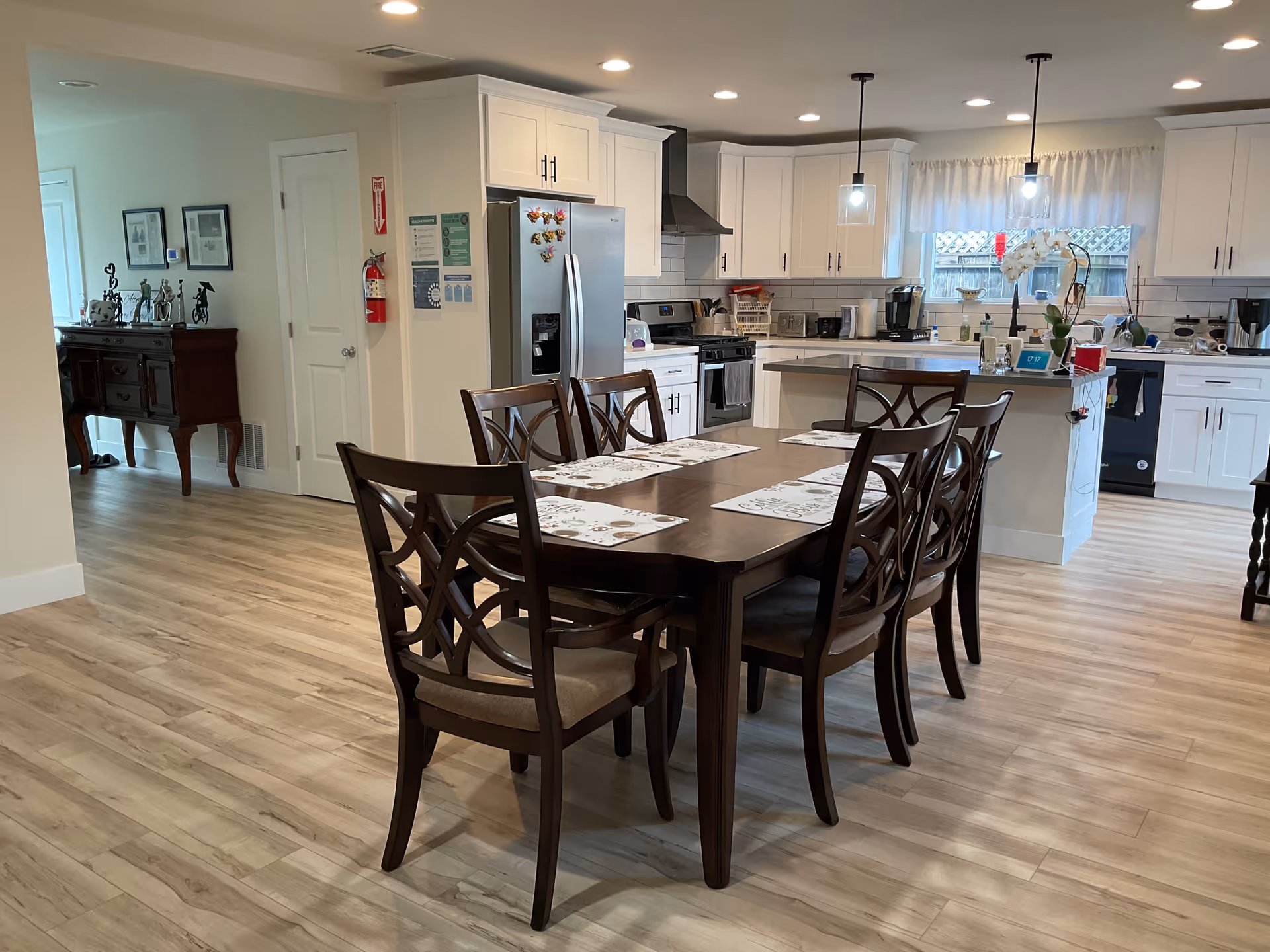 A bright and spacious kitchen and dining area with light wood flooring. The dining table is dark wood with six matching chairs, each with a placemat. The kitchen has white cabinets, stainless steel refrigerator, stove, and dishwasher. Two pendant lights hang over the kitchen island, which has a vase with white flowers. A window with sheer curtains lets in natural light.