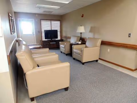 A senior living lounge with beige recliners, a TV, lamp, and entry doorway.