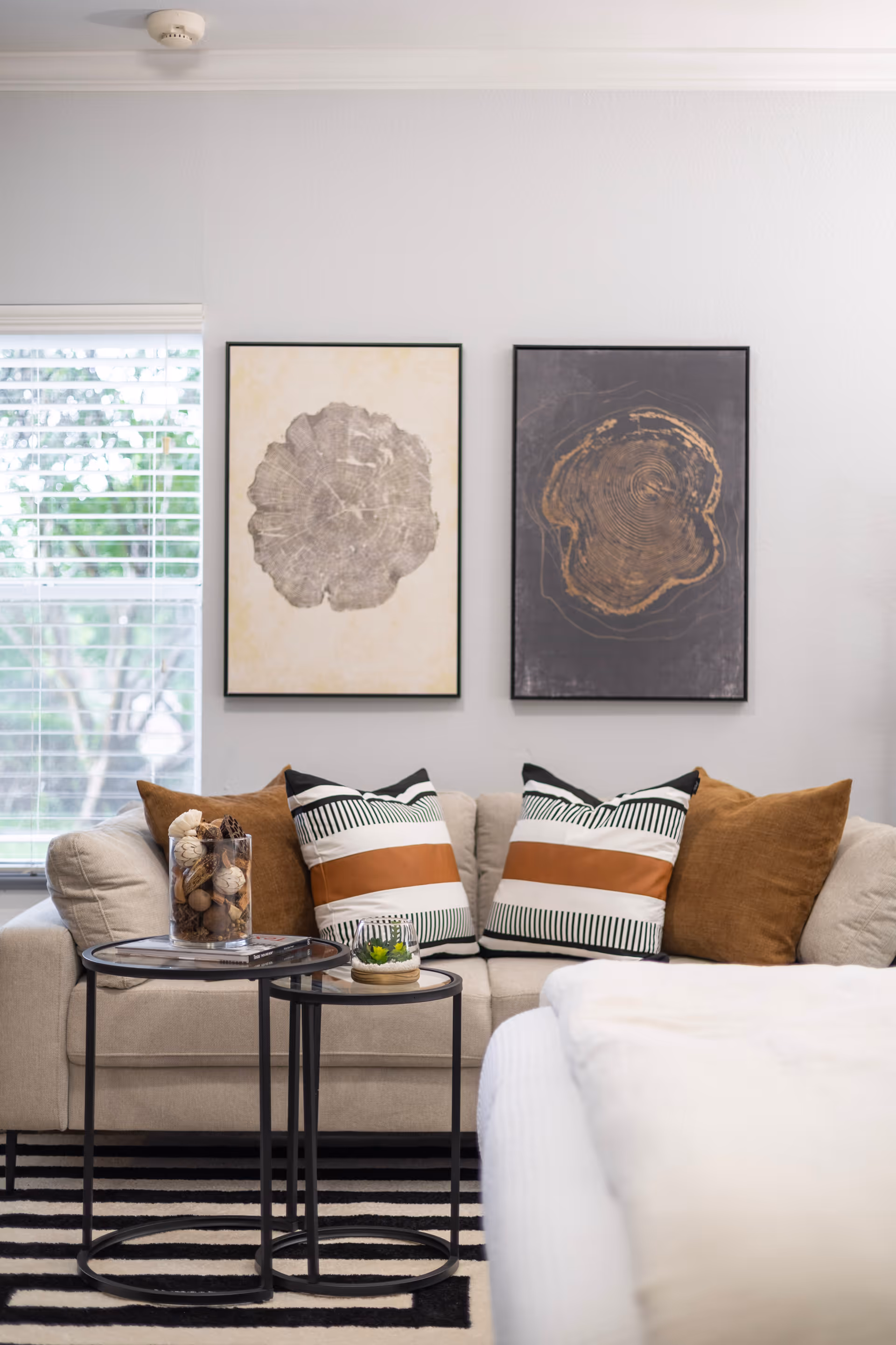 A cozy living room area with a beige sofa adorned with four decorative pillows, two with black and white stripes and brown bands, and two solid brown pillows. In front of the sofa are two round nesting tables with black metal frames, one holding a glass container filled with decorative natural elements and the other a small plant. Behind the sofa, two framed artworks depicting tree rings hang on a light gray wall. A window with white blinds is partially visible on the left side.