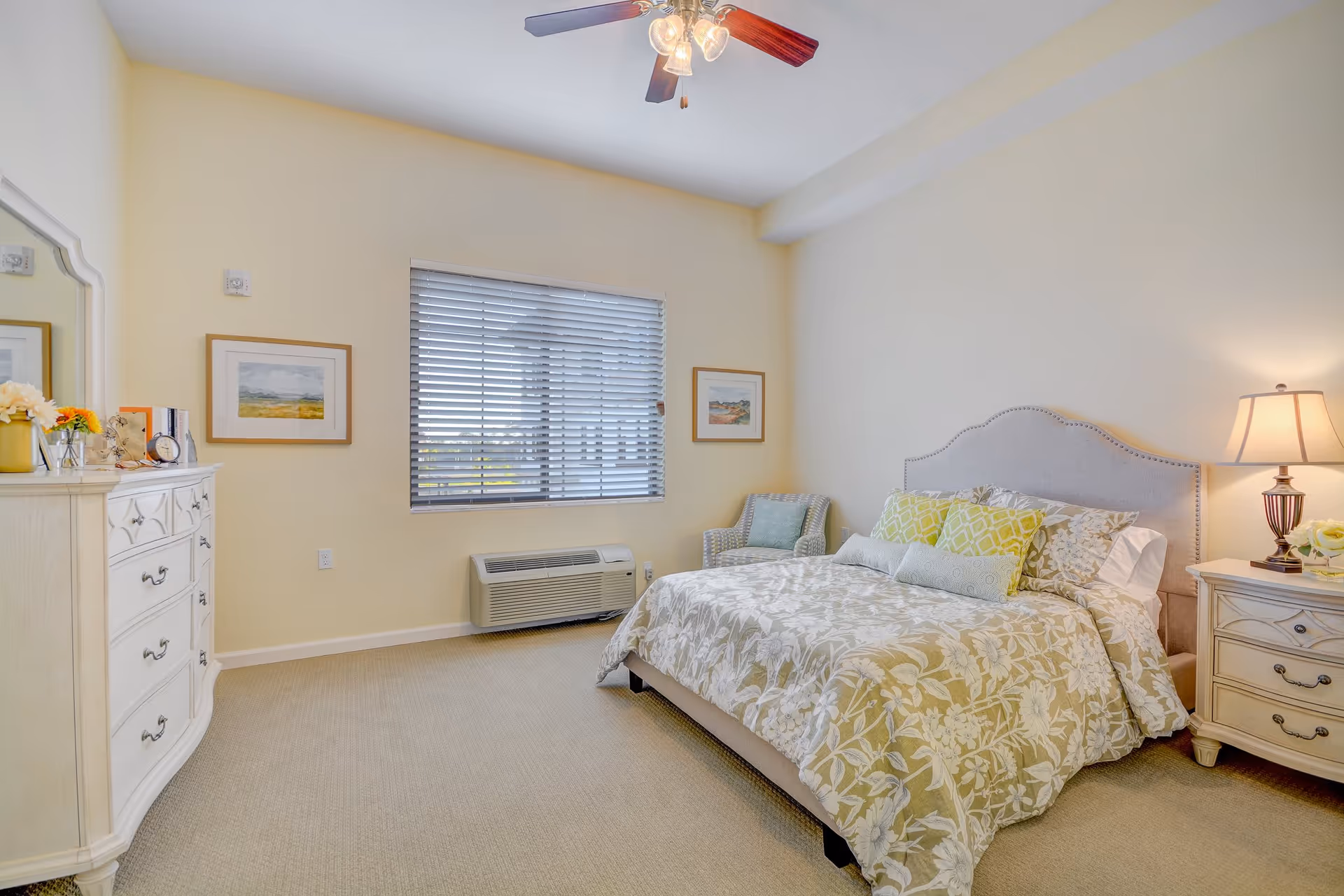 A bright and cozy bedroom with a large bed featuring floral bedding and multiple pillows. There is a cushioned armchair in the corner near a window with blinds. A ceiling fan with lights is mounted on the ceiling. The room also includes a white dresser with decorative items and a nightstand with a lamp and flowers.