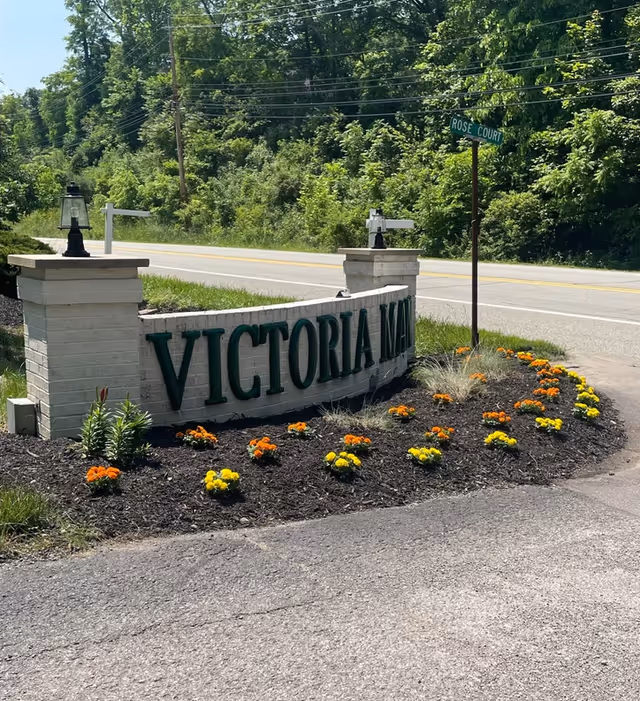 Stone entrance sign reading 'Victoria Manor' with a landscaped flowerbed by a roadside and trees in the background.