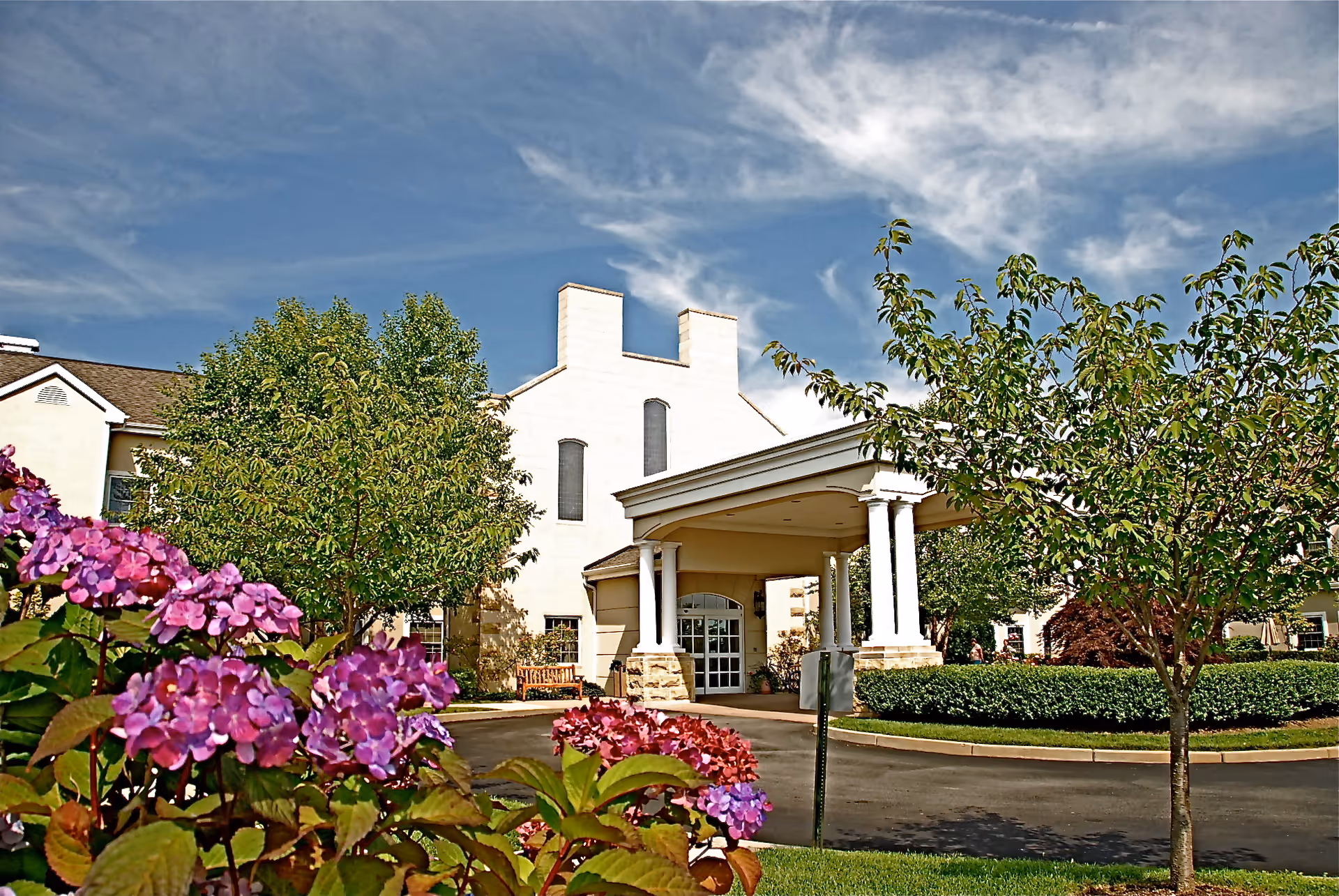 Front entrance of a nursing and rehabilitation center with a covered porte-cochère, landscaped trees and purple flowers under a blue sky.
