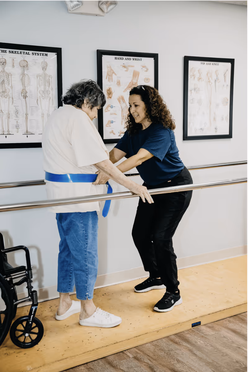 A physical therapist assists an elderly woman with walking exercises using parallel bars in a rehabilitation room. The elderly woman is wearing a blue gait belt and casual clothes, while the therapist is dressed in a navy shirt and black pants. Anatomical posters of the skeletal system, hand and wrist, and hip and knee are visible on the wall behind them. A wheelchair is positioned nearby.