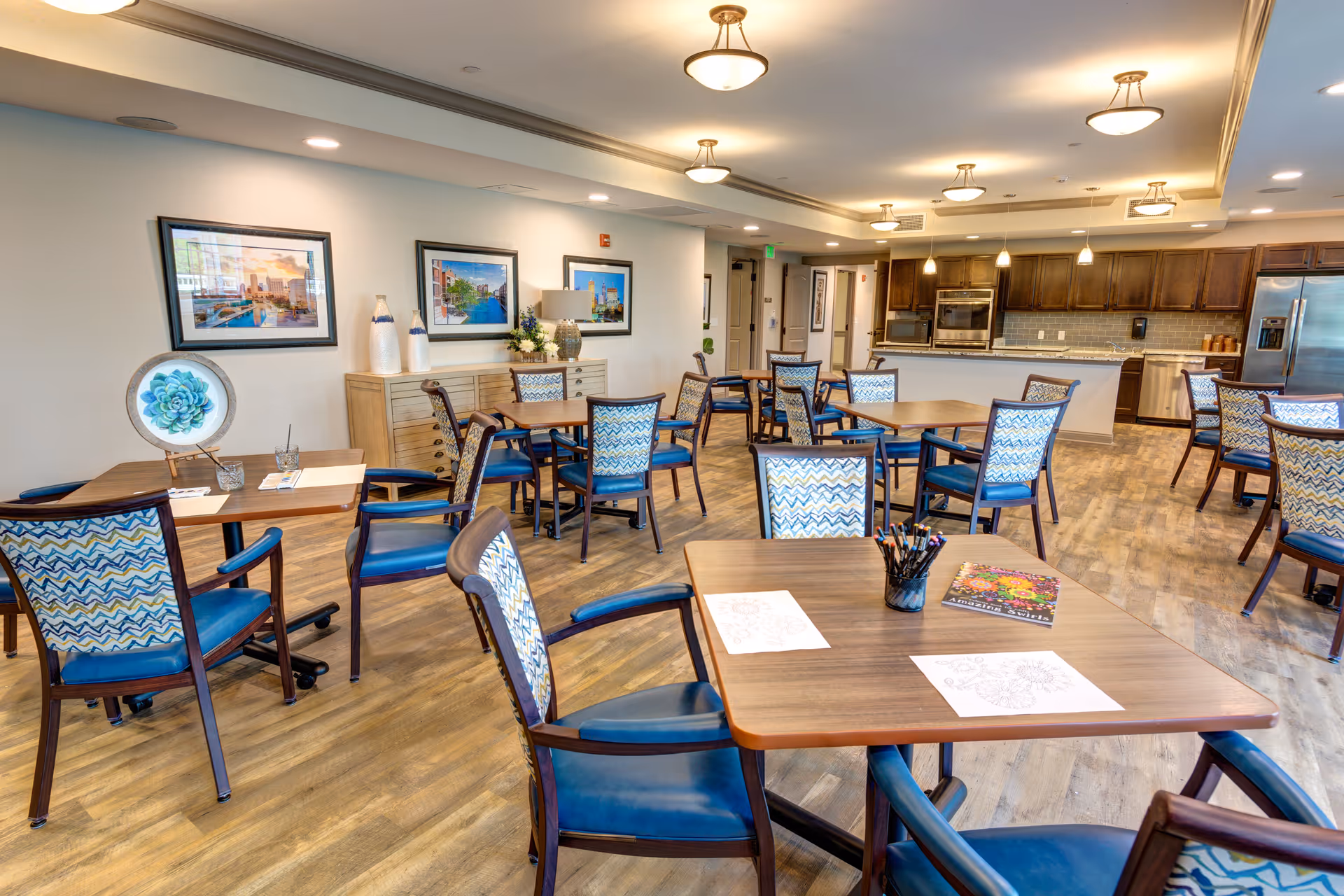 A bright and spacious common area with several wooden tables and chairs featuring blue patterned upholstery. The room has wood flooring, framed artwork on the walls, and a modern kitchen area with stainless steel appliances in the background. The tables have coloring sheets and colored pencils on them, suggesting an activity space.