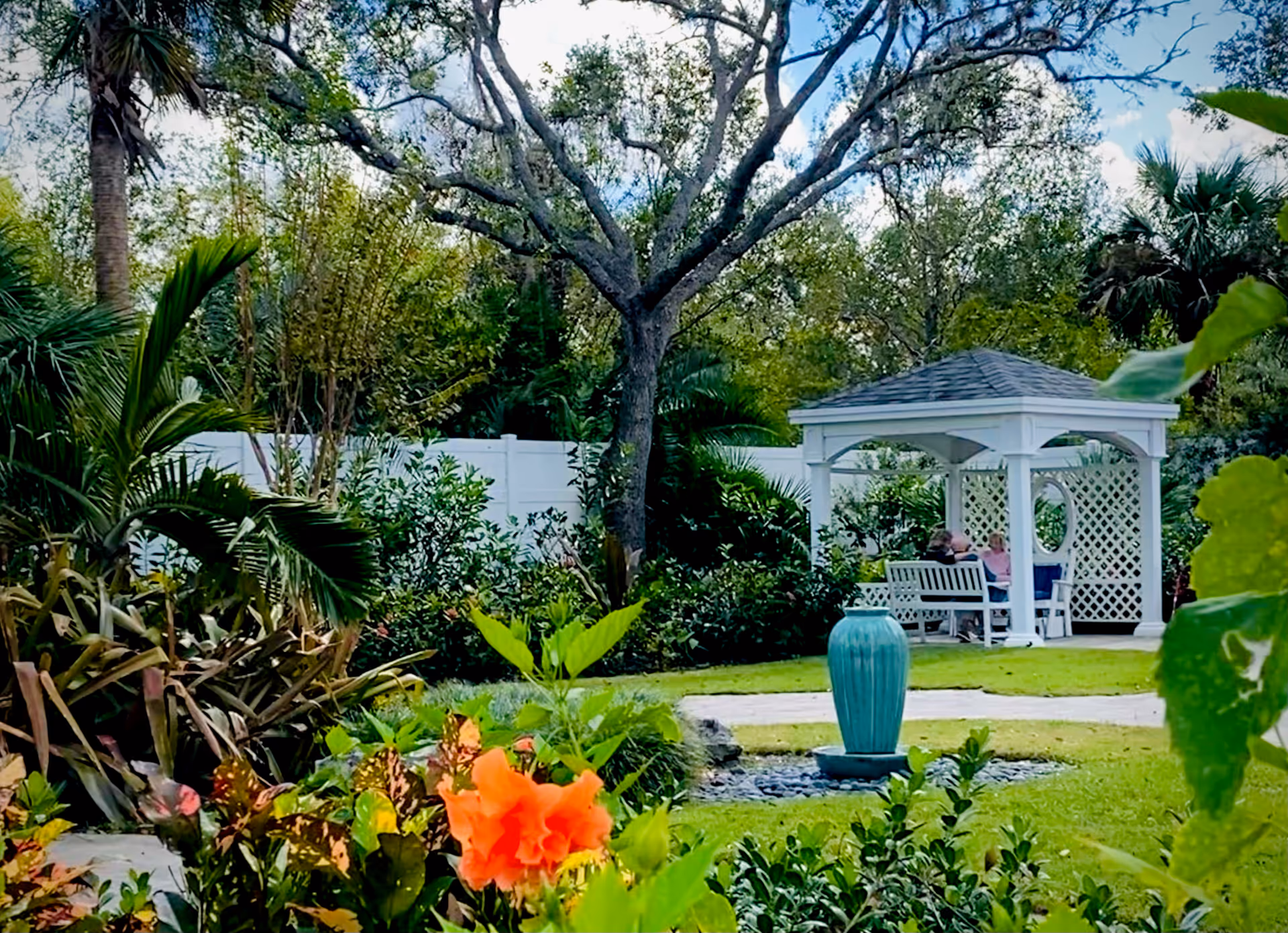 A lush garden area with various green plants and an orange flower in the foreground. In the background, there is a white gazebo with lattice sides where two people are seated on a bench. A blue ceramic water fountain is also visible on the grass near a paved pathway. Tall trees and a white fence surround the garden.