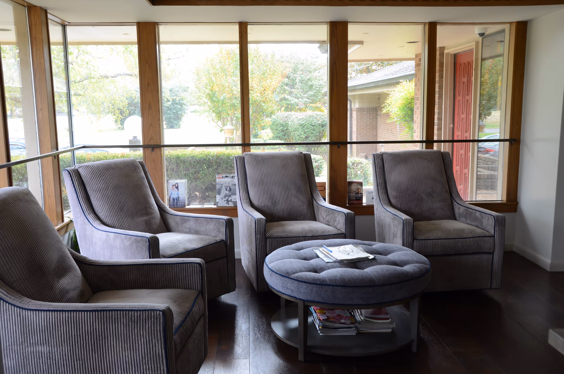 A bright sitting area with four gray upholstered armchairs arranged around a round tufted ottoman in front of large wood-framed windows.