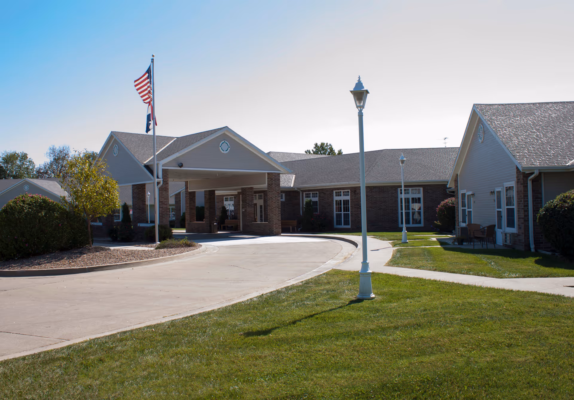 Exterior view of Country Club Rehab & Healthcare Center showing a single-story building with a covered entrance, two flagpoles with American and state flags, a curved driveway, green lawn, and lamp posts.