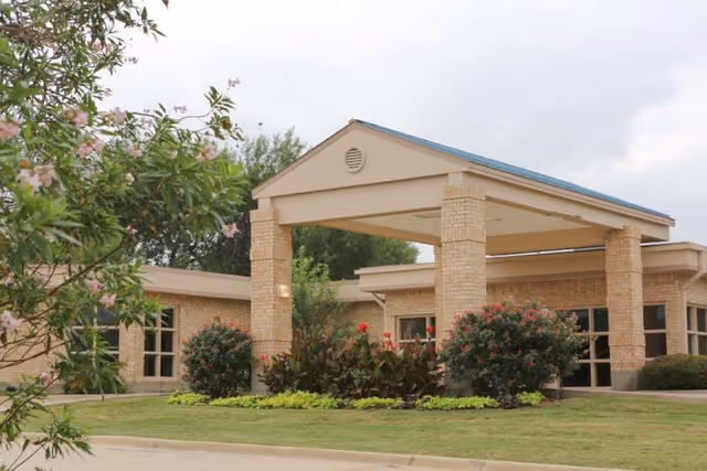 Exterior view of a single-story brick building with a covered entrance supported by four brick columns. The building is surrounded by green grass, shrubs, and flowering plants under a cloudy sky.