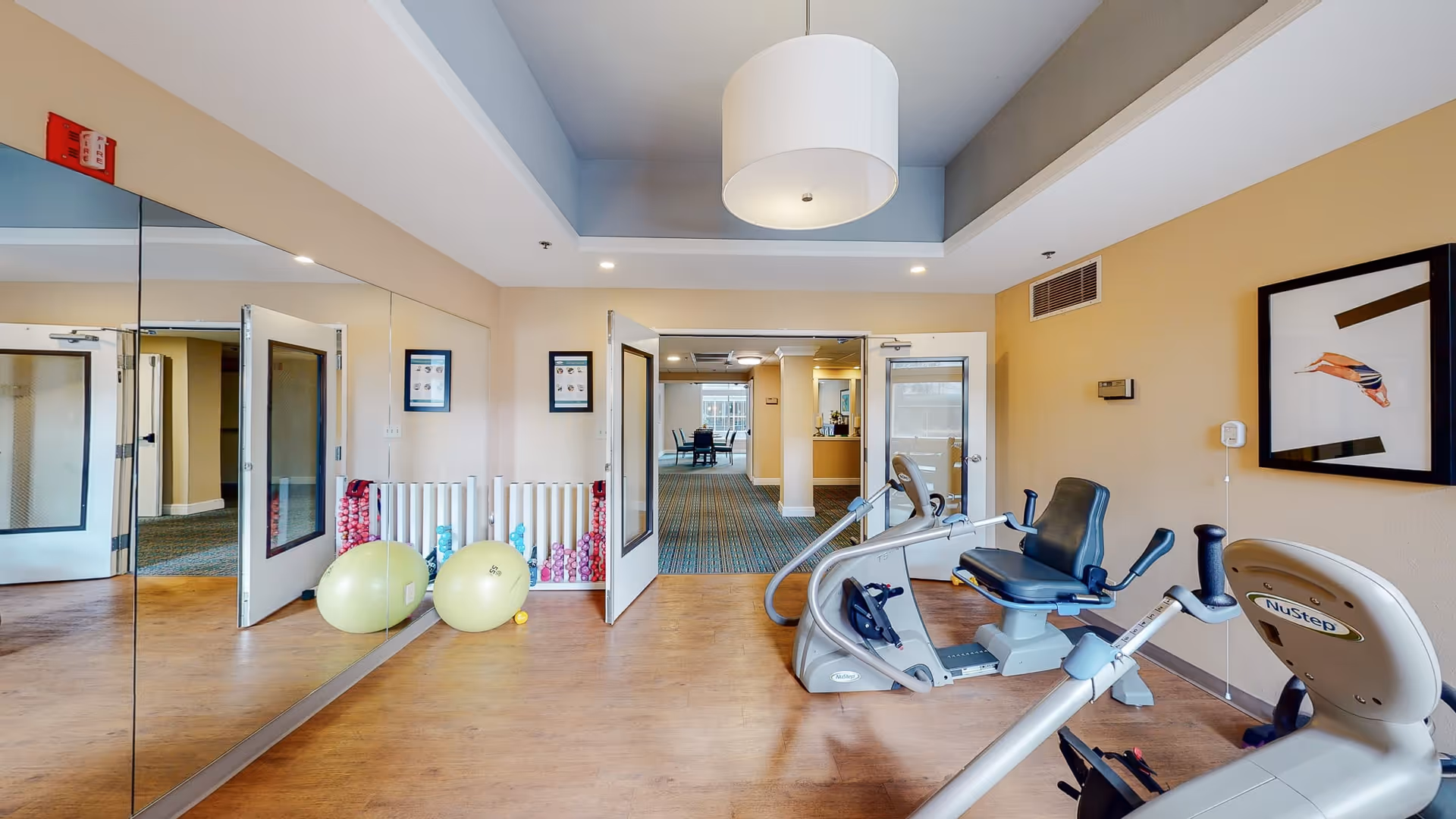 A small exercise room with wooden flooring featuring two recumbent exercise bikes, two large yellow exercise balls, and a rack of colorful dumbbells against a mirrored wall. The room has beige walls, a large ceiling light fixture, and a framed picture of a diver on the right wall. Double glass doors lead to a carpeted hallway with seating visible in the distance.