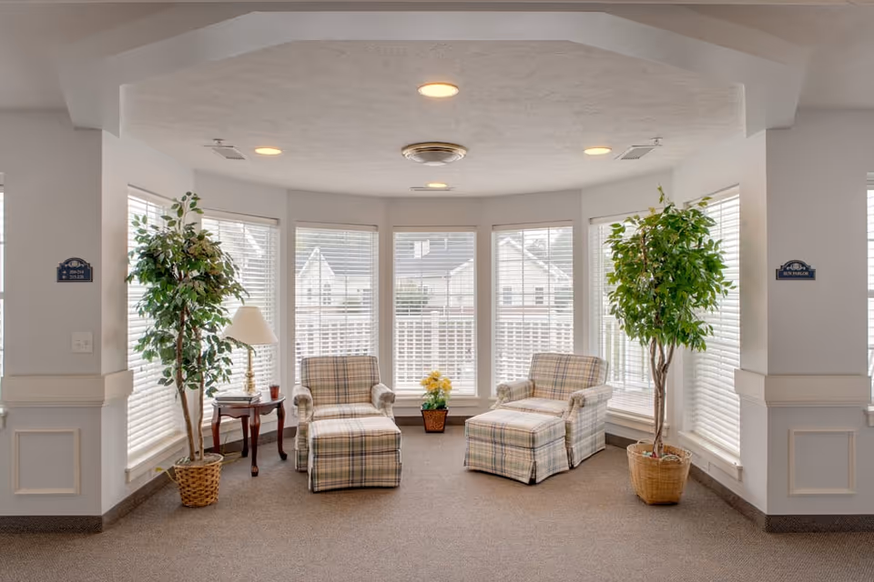 A cozy sitting area with two plaid armchairs and matching ottomans placed in front of large windows with white blinds. There are two potted plants on either side and a small wooden side table with a lamp and a cup. The space has a neutral color palette with beige carpet and white walls.