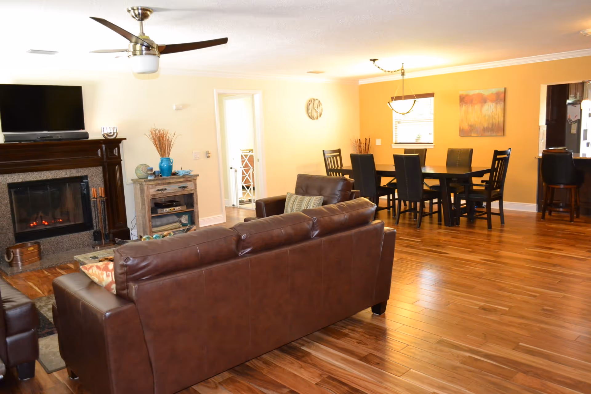 Bright open living and dining area with a brown leather sofa, fireplace, hardwood floors, and a dining table in the background.