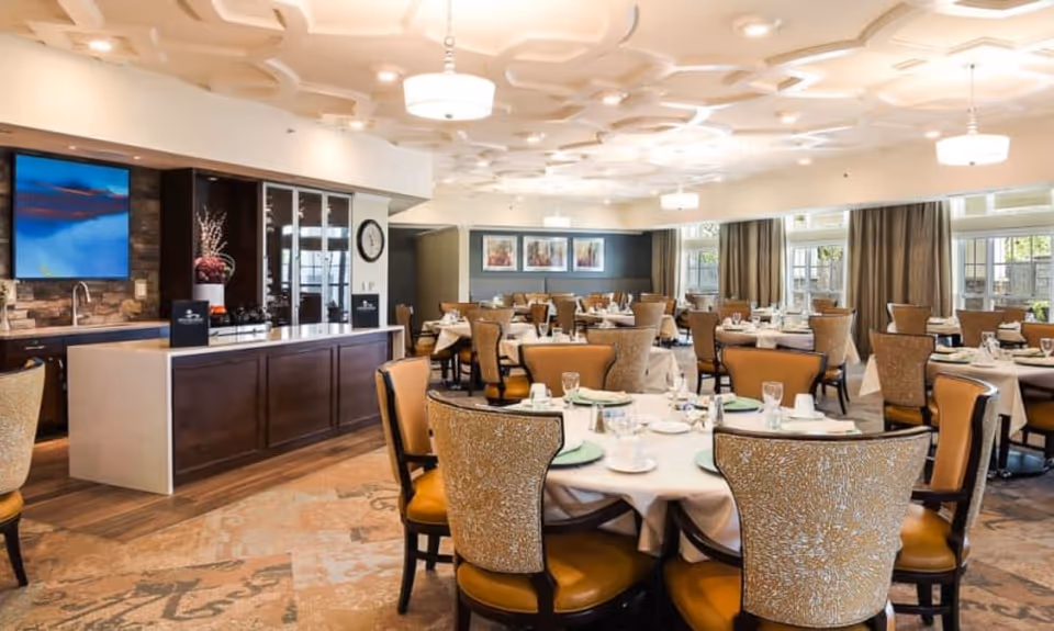 Spacious, well-lit dining room with round tables set with plates, glasses, and upholstered chairs.