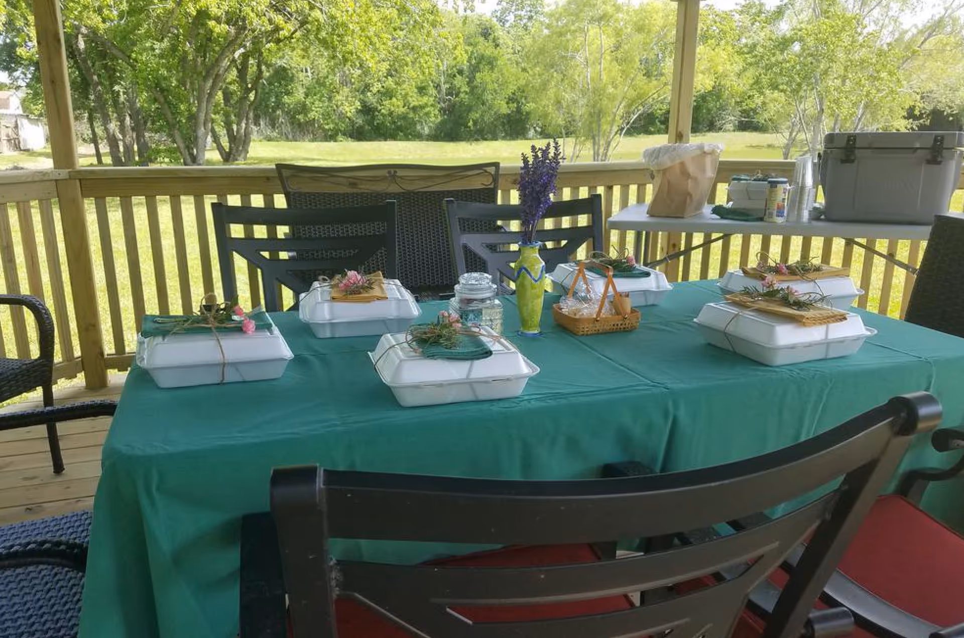 Outdoor covered patio area with a table covered in a green tablecloth. On the table are six white takeout containers, each decorated with a green napkin, twine, and small flowers. There is a green vase with purple flowers in the center and a small basket with utensils. The background shows a grassy yard with trees and a wooden railing around the patio.
