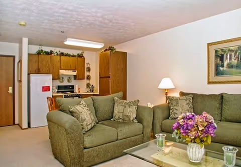 Interior view of a living room and kitchen area in a senior living facility. The living room features two green upholstered sofas with patterned cushions, a glass coffee table with a floral arrangement and two candle holders, a table lamp, and a framed painting on the wall. The kitchen area has wooden cabinets, a white refrigerator, a stove, and decorative plates on the wall above the stove.