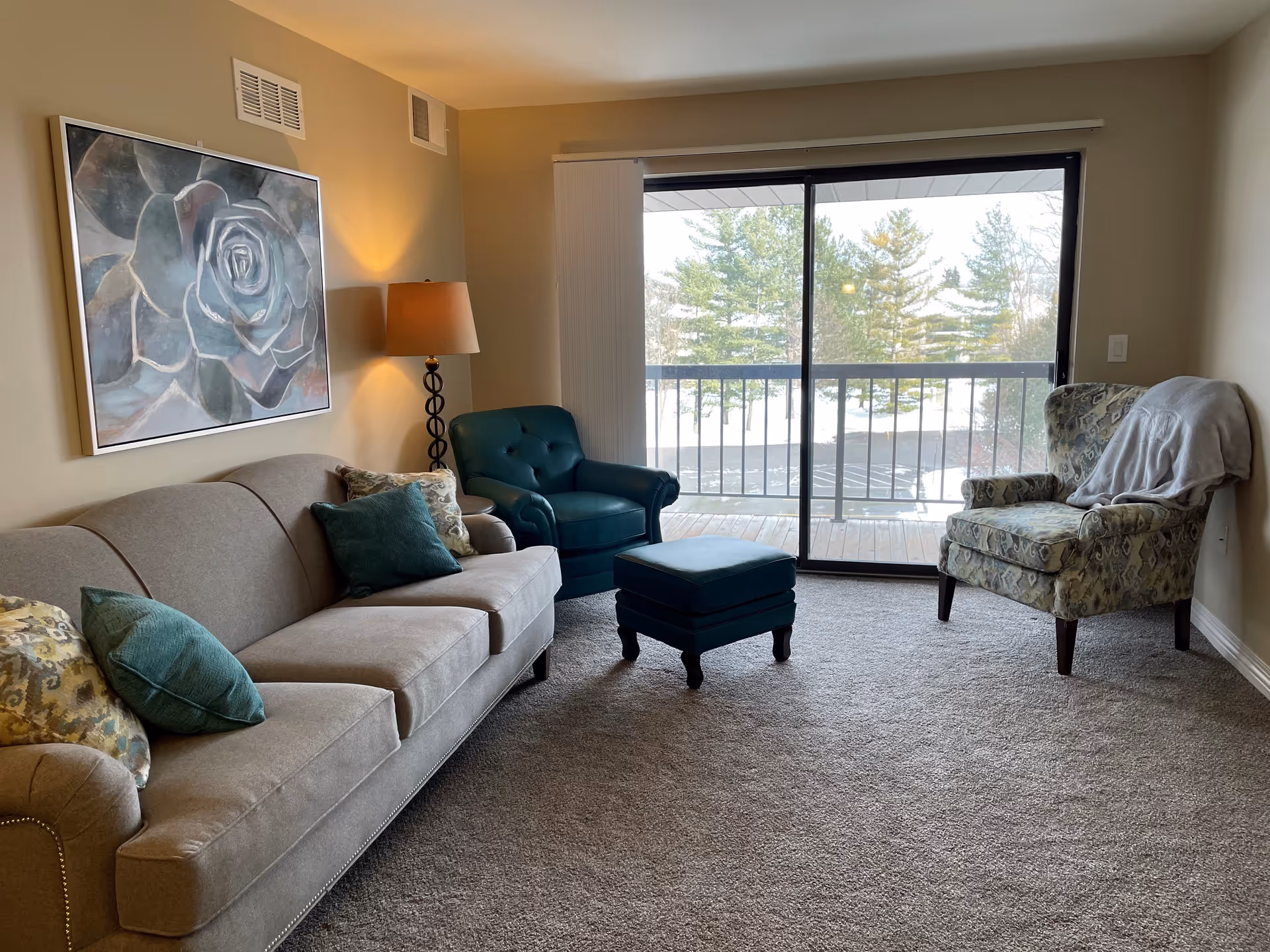 A cozy living room with a sofa, two armchairs, an ottoman, a floor lamp, wall artwork, and a sliding glass door leading to a balcony.