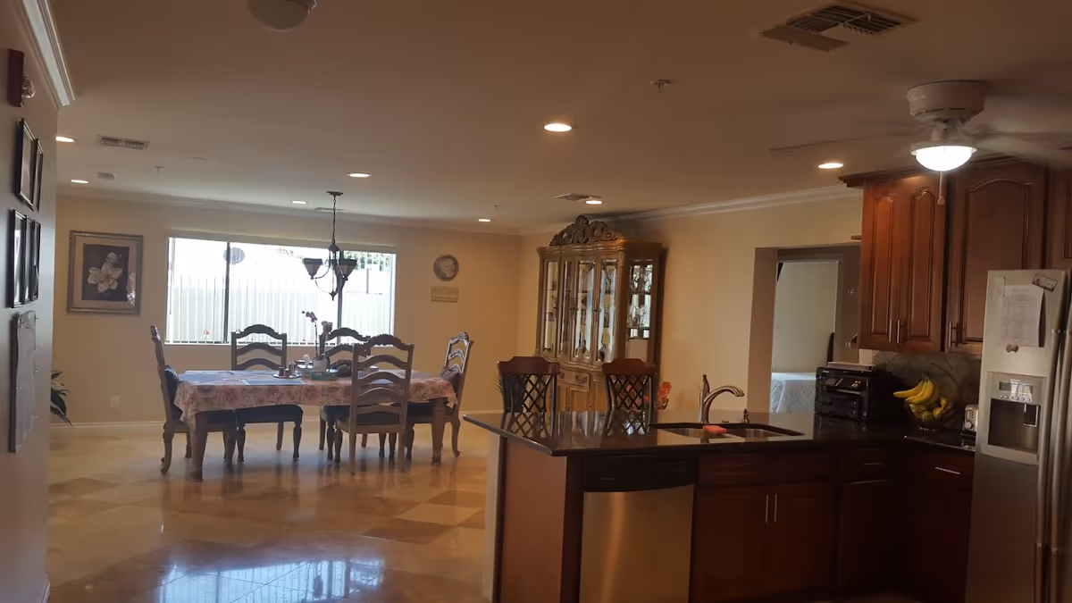 Interior view of a dining area and kitchen in an assisted living facility. The dining area features a table with six chairs and a floral tablecloth, a chandelier hanging above, a large window letting in natural light, and a wooden china cabinet against the wall. The kitchen has dark granite countertops, wooden cabinets, a stainless steel refrigerator, a dishwasher, and a ceiling fan with a light.