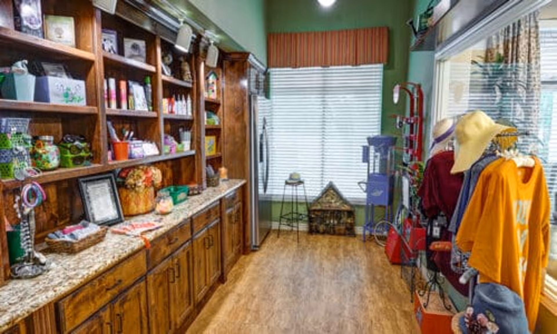 Interior view of a small shop or boutique area inside a facility, featuring wooden shelves stocked with various items such as jars, bottles, and decorative pieces on the left side. On the right side, there are racks displaying clothing and hats near a large window with blinds. The floor is wooden, and the walls are painted green.