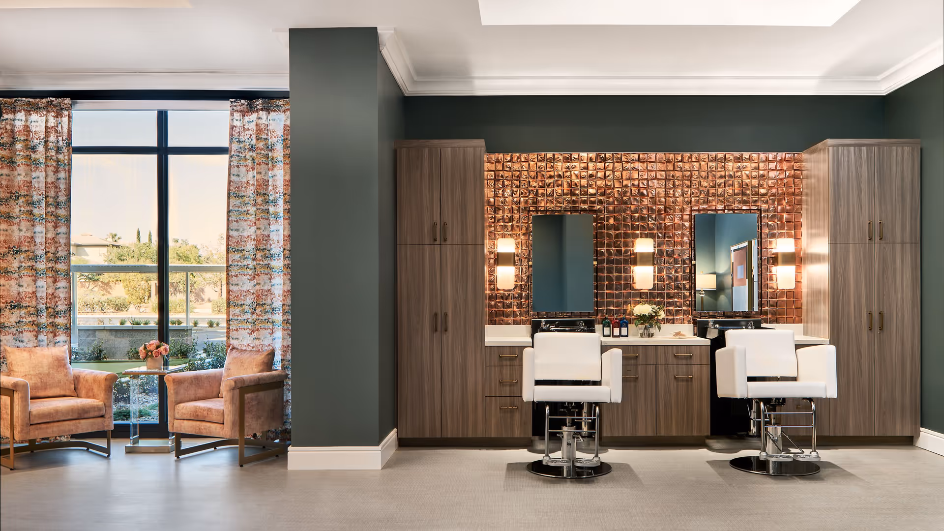 A stylish salon area with two white salon chairs in front of a copper-tiled wall with two mirrors and wall-mounted lights. On either side of the mirrors are tall wooden cabinets. To the left, there is a large window with floral curtains and two pink upholstered armchairs with a small glass table between them holding a vase of flowers.
