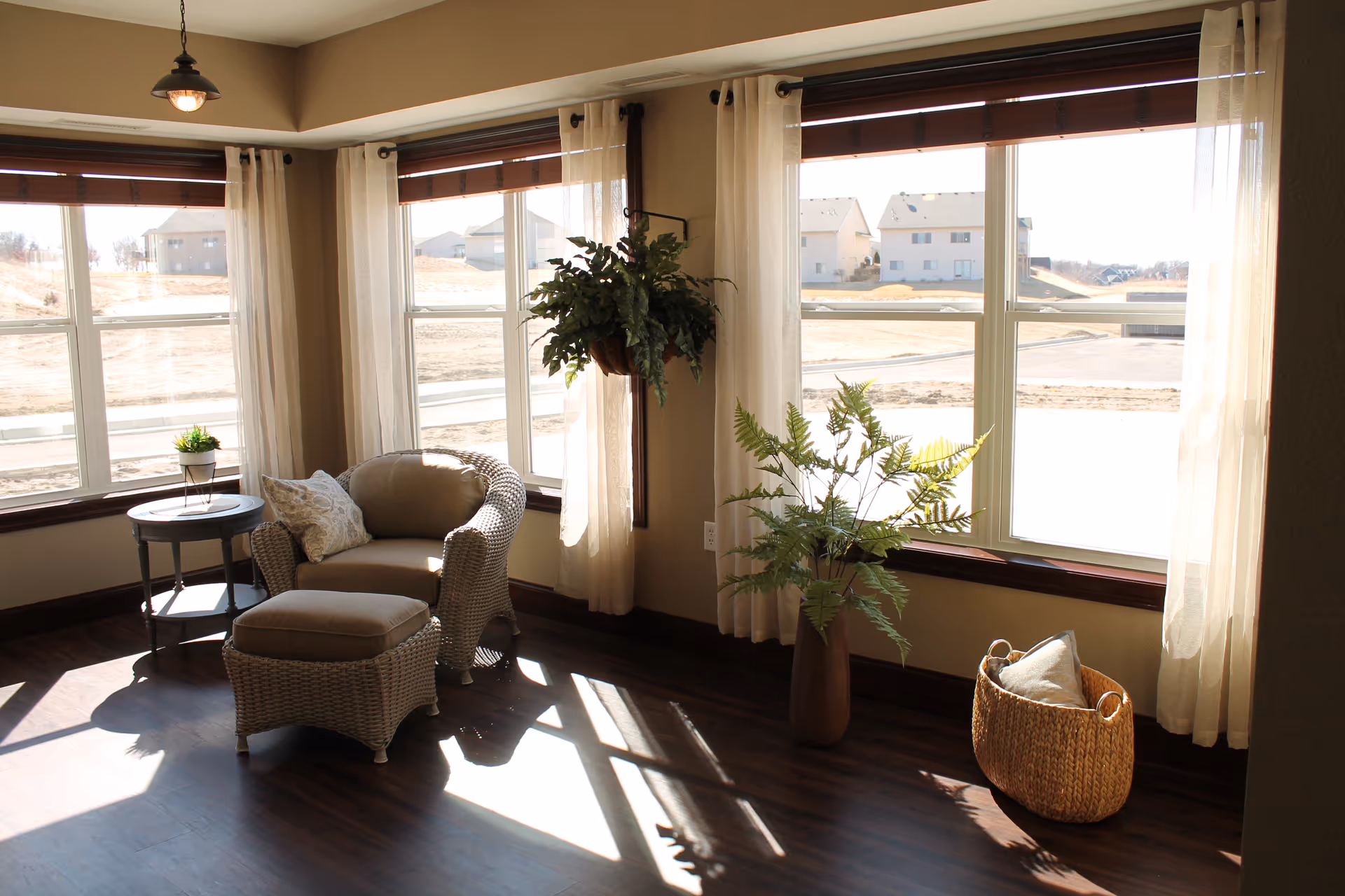 A cozy corner of a living room with large windows letting in natural light. There is a wicker armchair with a matching ottoman, a small round side table with a small plant, a hanging potted plant, a tall vase with green foliage, and a woven basket with a pillow inside. Light curtains frame the windows, and the floor is dark wood.