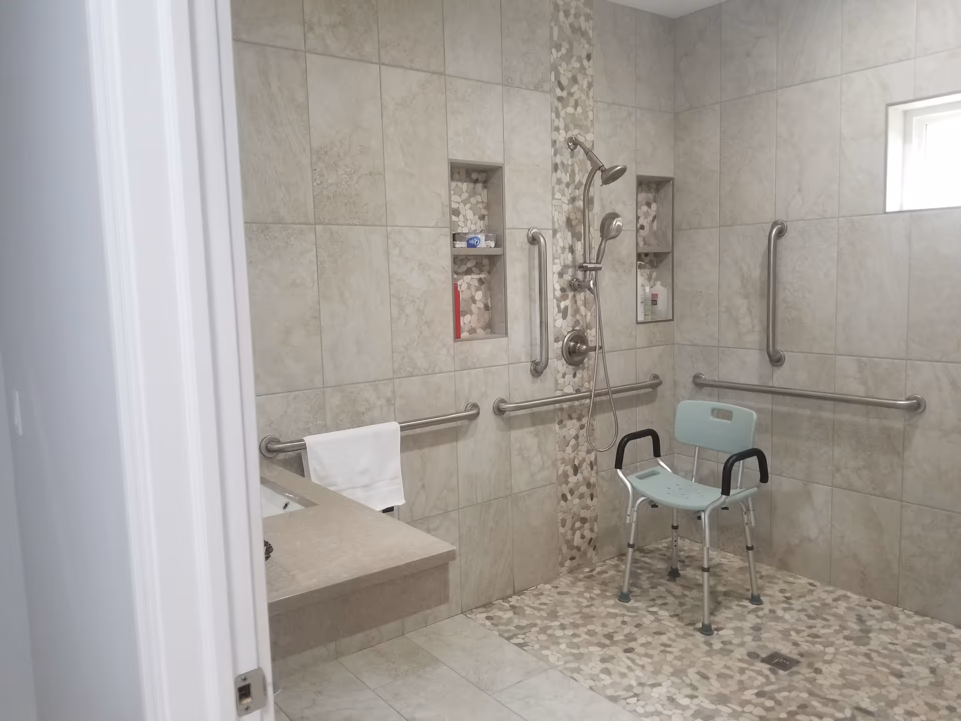 Accessible shower area with beige tiled walls and pebble-patterned floor and accent strip. The shower has a handheld showerhead, multiple grab bars, built-in shelves with toiletries, and a light blue shower chair with armrests. A towel hangs on a grab bar near a beige countertop with a sink.