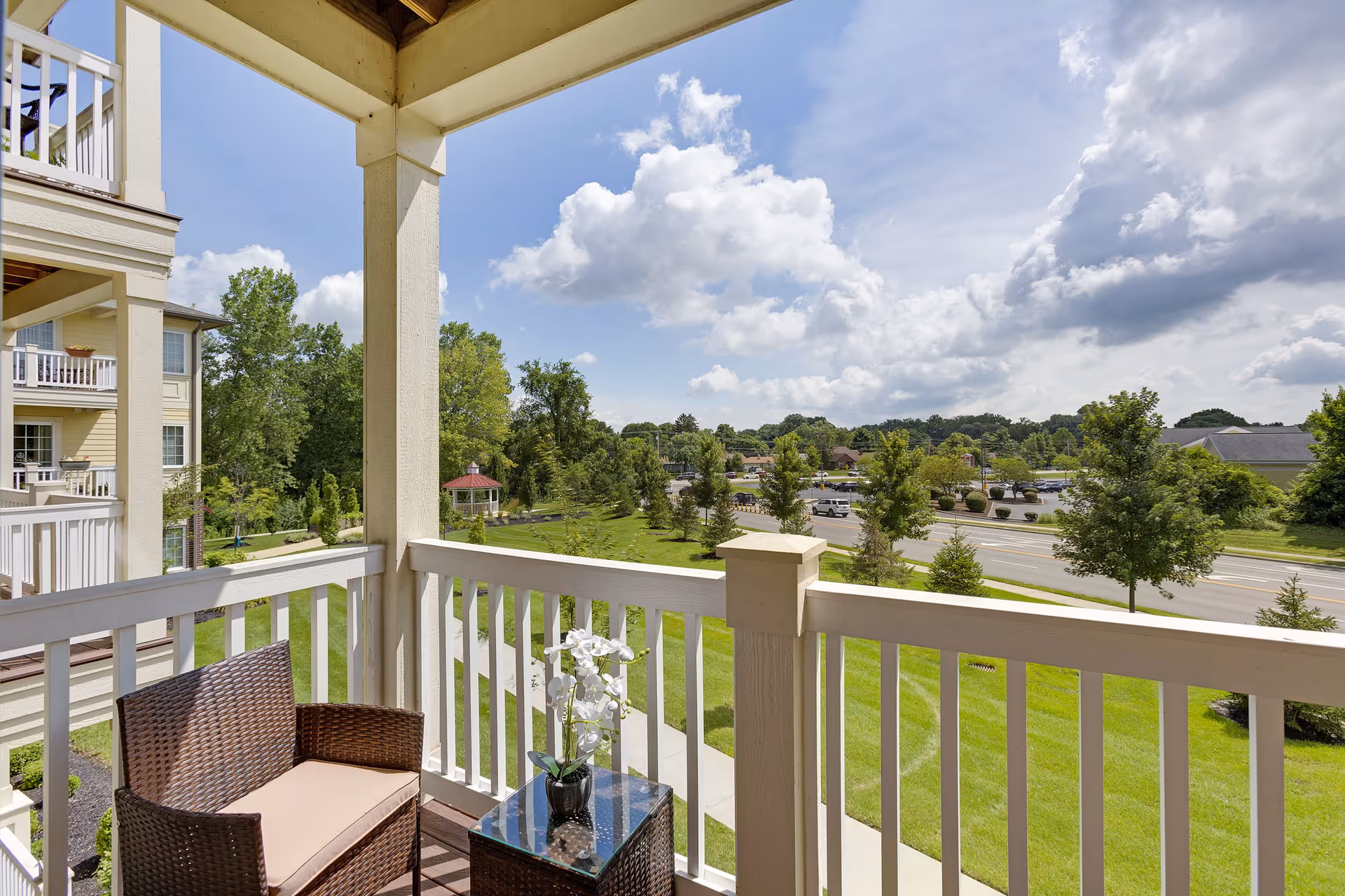 View from a covered balcony with a wicker chair and a small table holding a white orchid plant. The balcony overlooks a green lawn, trees, a gazebo, a road, and a parking lot under a partly cloudy sky.