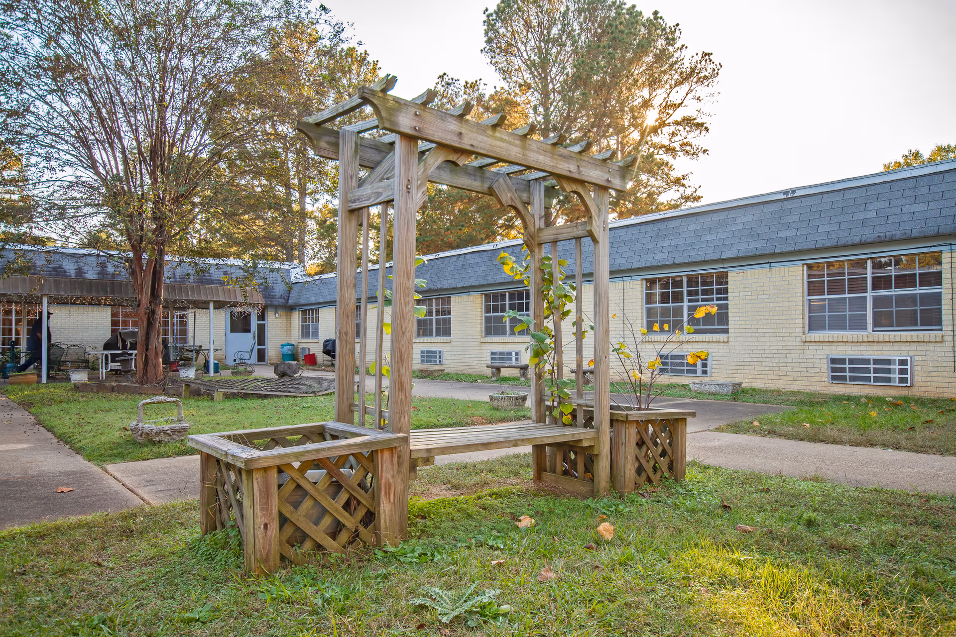 Outdoor courtyard area of a senior living facility with a wooden pergola bench surrounded by grass and paved walkways. The building in the background has light yellow brick walls and multiple windows with air conditioning units beneath them. Trees with autumn foliage are visible around the courtyard.