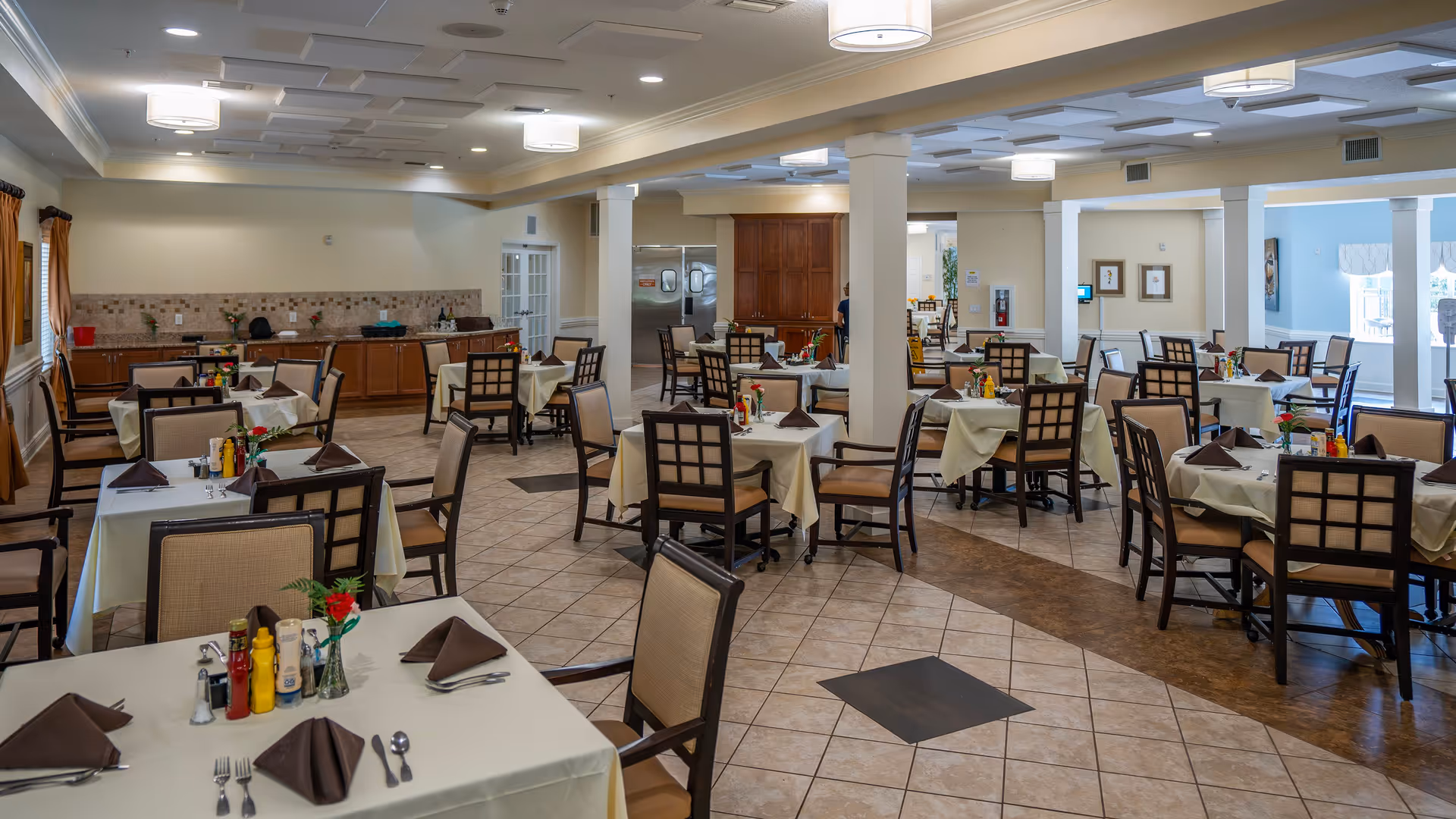 A spacious dining room with multiple tables covered in white tablecloths, each set with brown napkins, silverware, condiments, and small flower vases. The room has tiled floors, beige walls, and ceiling lights. There are wooden chairs with cushioned seats around the tables, and a kitchen area with cabinets and stainless steel appliances is visible in the background.