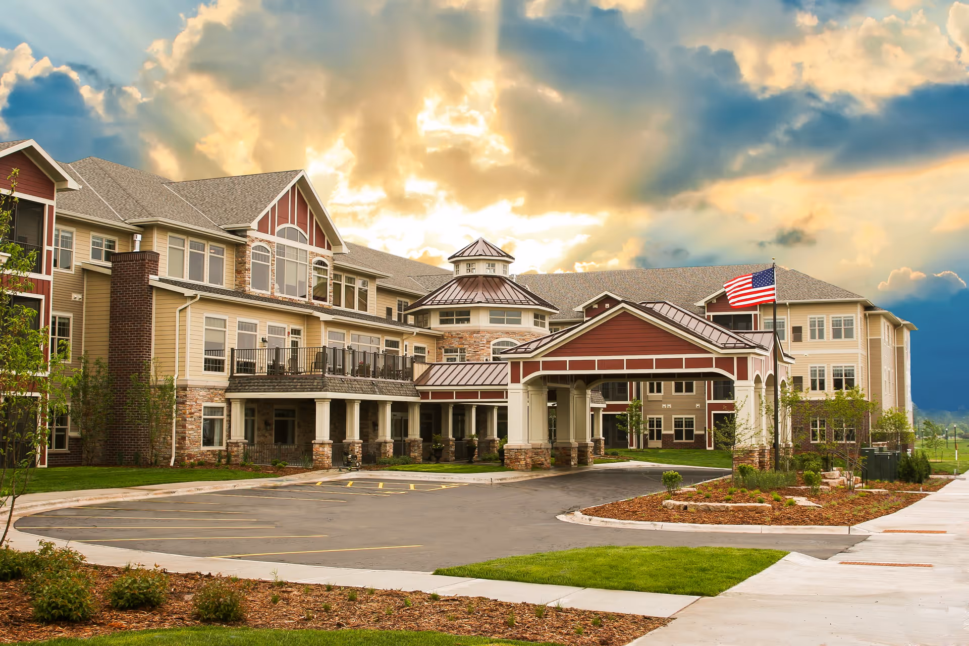 Exterior view of a large senior living facility building with a covered entrance, multiple windows, and an American flag on a flagpole. The sky is partly cloudy with sunlight breaking through.