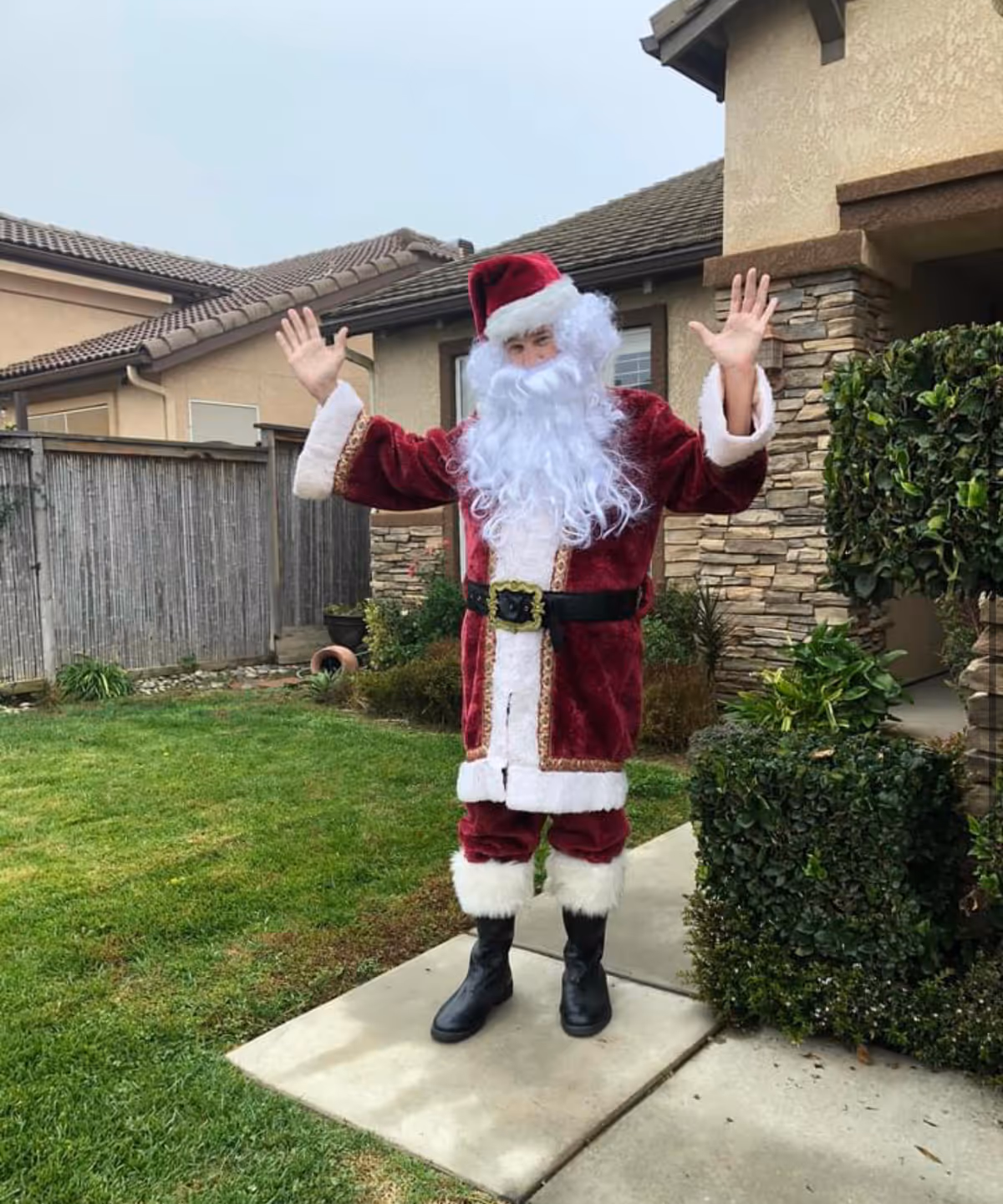 A person dressed in a Santa Claus costume with a red suit, white beard, and black boots stands on a concrete pathway in front of a residential-style building with stone and stucco exterior. The person has both hands raised and is standing near green bushes and a grassy lawn.