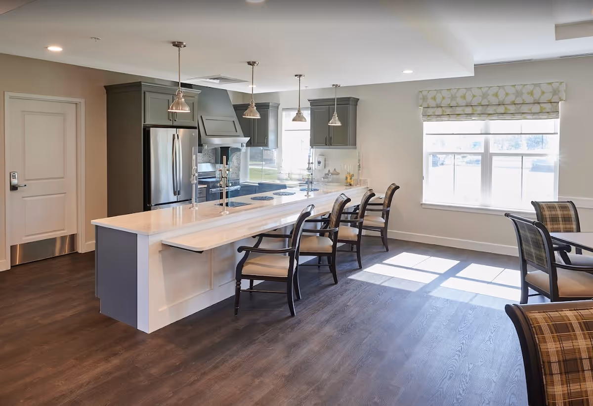 A bright, modern kitchen area with a long white countertop island featuring four wooden chairs with cushioned seats. The kitchen has gray cabinets, stainless steel refrigerator, and three pendant lights hanging above the island. There is a large window with patterned blinds letting in natural light, and a round table with plaid cushioned chairs is partially visible on the right side.