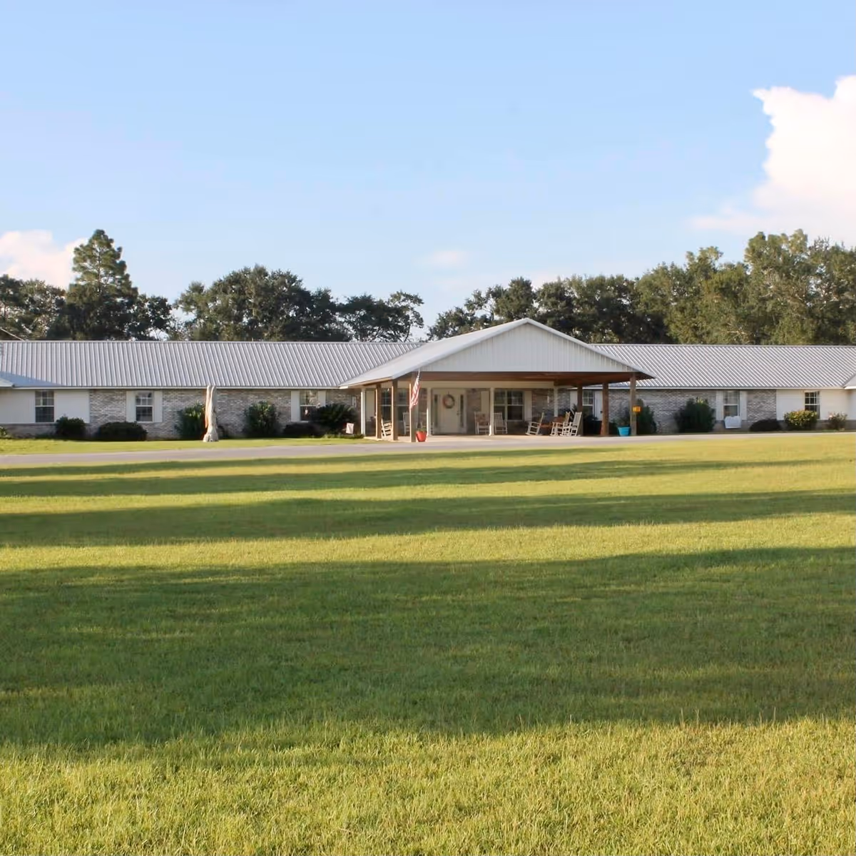 Single-story building with a metal roof and a covered porch area with rocking chairs, surrounded by a large green lawn and trees in the background under a clear blue sky.