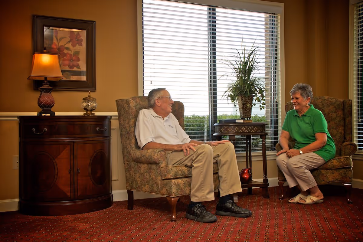 An elderly man and woman sitting in upholstered armchairs facing each other and smiling in a warmly lit room with a window covered by blinds behind them. There is a wooden side table with a decorative plant between them, and a wooden cabinet with a lamp and framed artwork on the wall to the left.