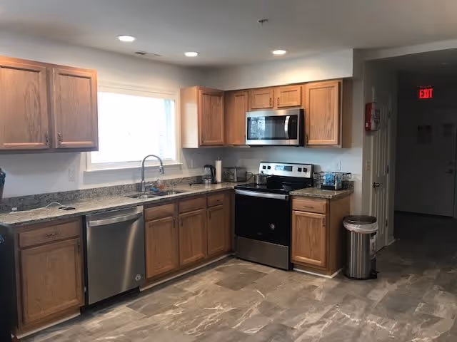 A clean and modern kitchen area with wooden cabinets, a stainless steel dishwasher, stove, microwave, and a sink under a window. The floor is tiled with a marble-like pattern, and there is a trash can near the entrance to a hallway with an illuminated exit sign.