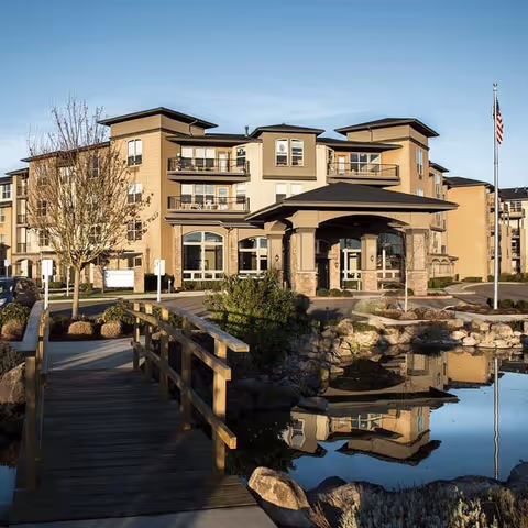 Exterior view of a multi-story senior living facility with a covered entrance, balconies, and large windows. In the foreground, there is a wooden footbridge over a pond with rocks and landscaping, and an American flag on a flagpole to the right.