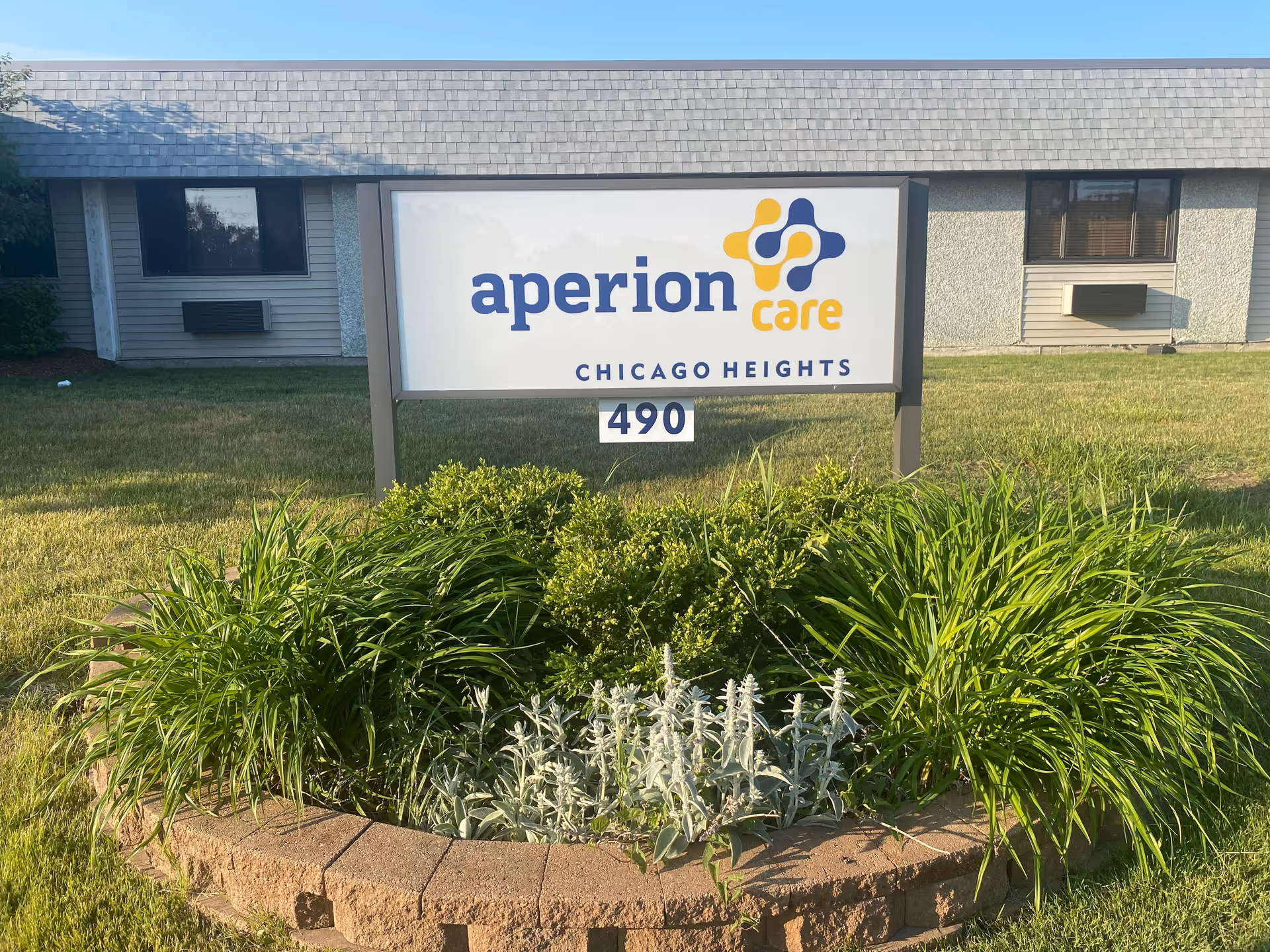 Outdoor view of a sign for Aperion Care Chicago Heights with the address number 490 displayed below. The sign is surrounded by a circular flower bed with green plants and shrubs, and a building with windows and air conditioning units is visible in the background.