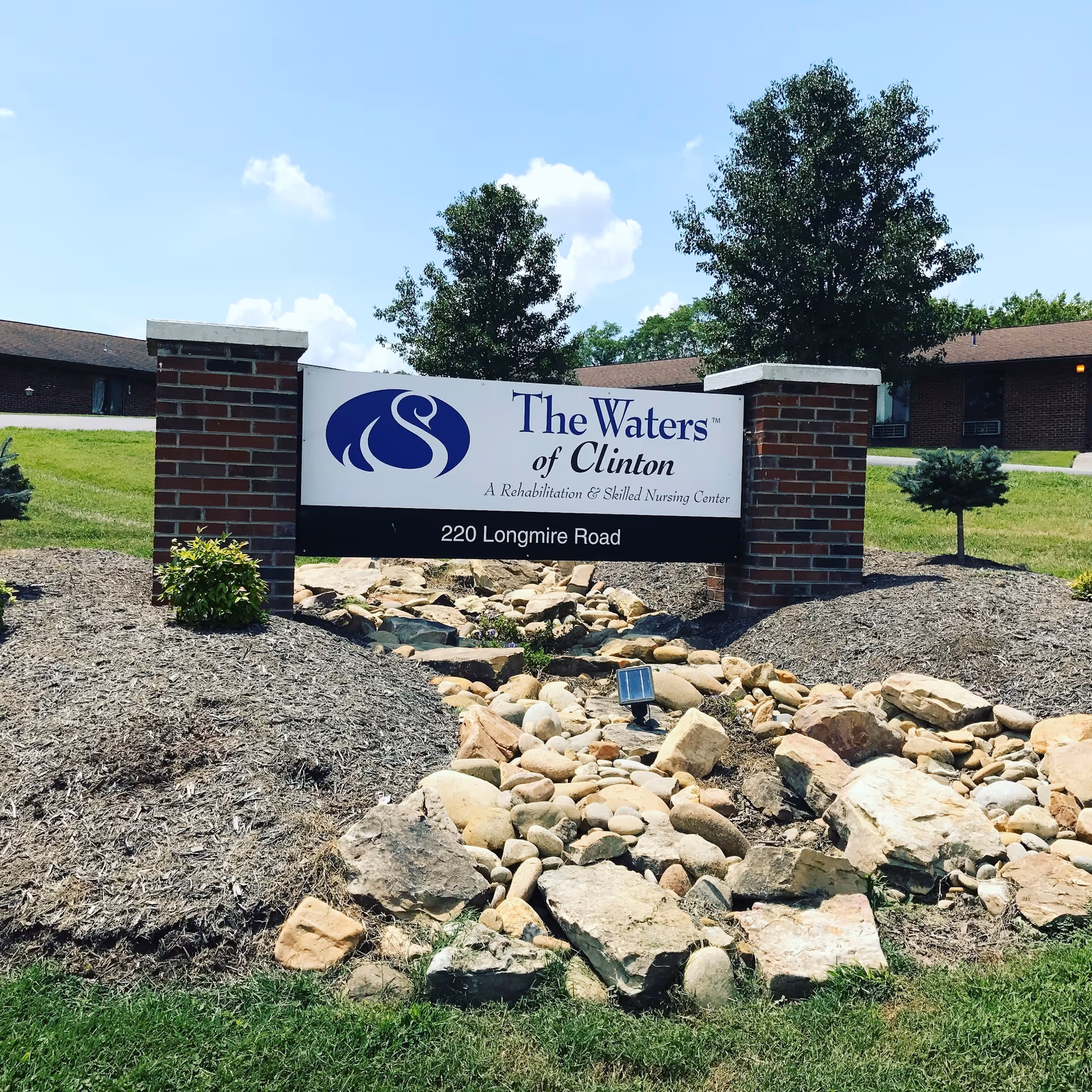 Front sign for The Waters of Clinton nursing center set between brick pillars with rock landscaping and the building behind.