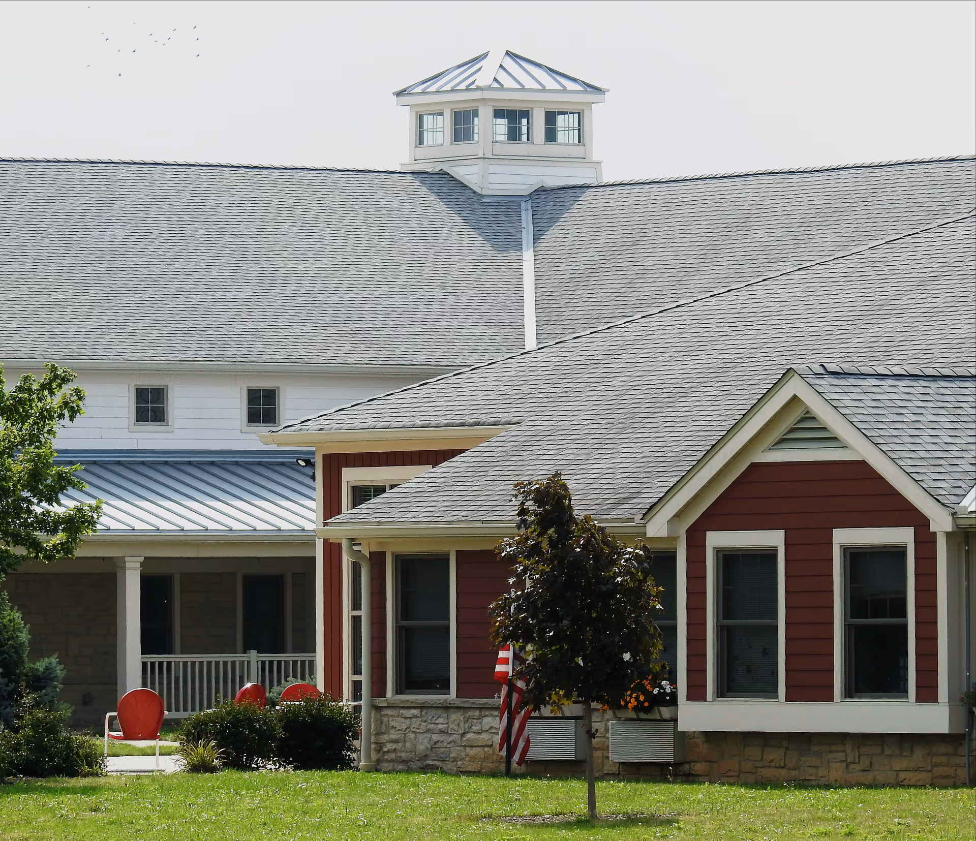 Exterior front of a red and white senior living building with multiple gabled roofs, a cupola, a porch with red chairs, and an American flag.