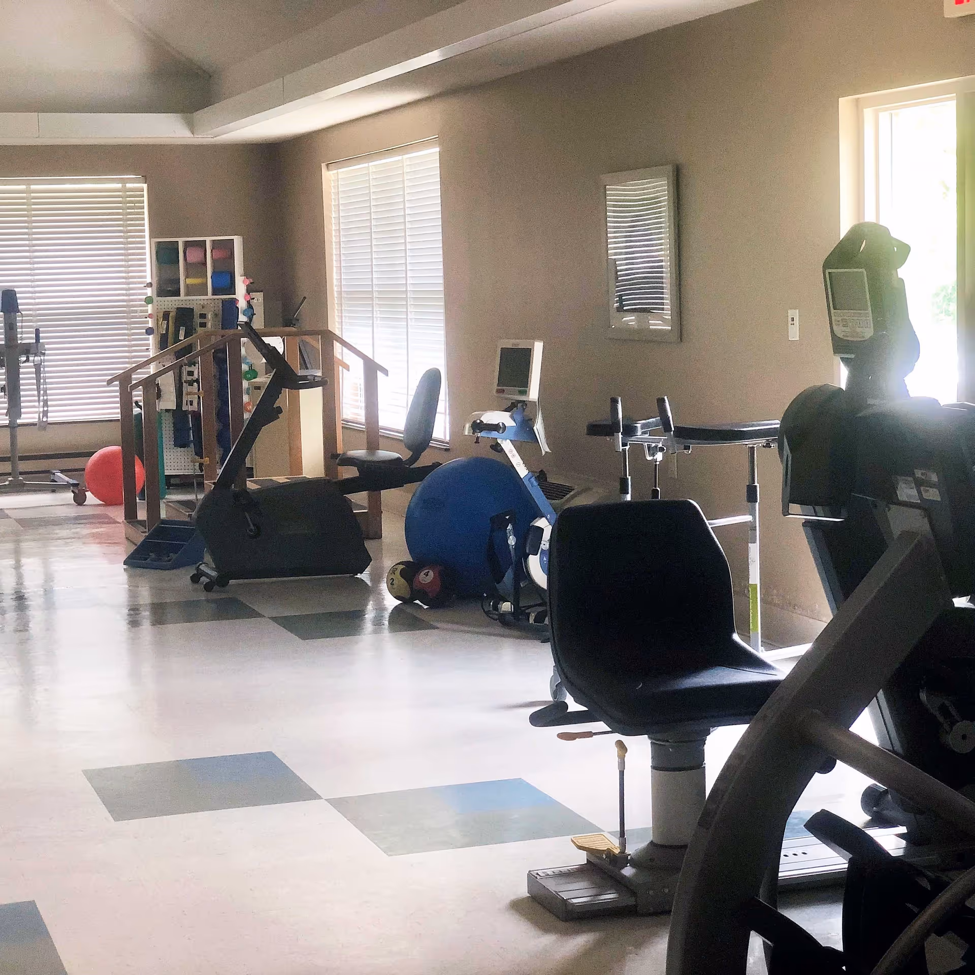 A rehabilitation exercise room with various fitness equipment including stationary bikes, a balance ball, parallel bars, and weights. The room has large windows with blinds allowing natural light to enter, and the floor has a pattern of square tiles in different shades.