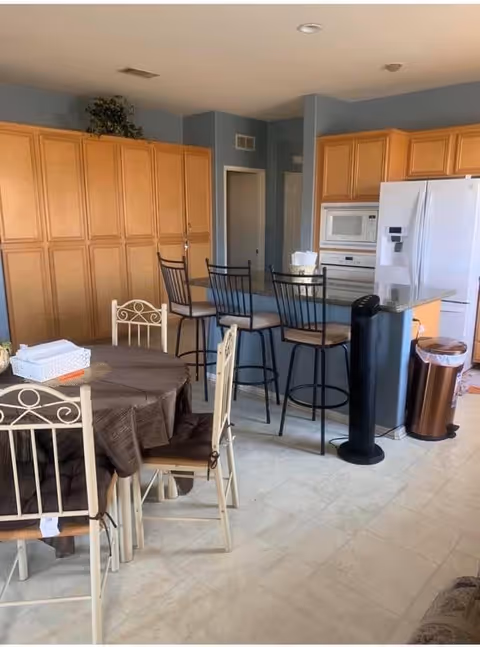 A kitchen and dining area with light wood cabinets, a white refrigerator, microwave, and oven. There is a round dining table with a brown tablecloth and four metal chairs with cushions. Three high bar stools are placed at the kitchen counter. A black tower fan and a copper-colored trash can are also visible.