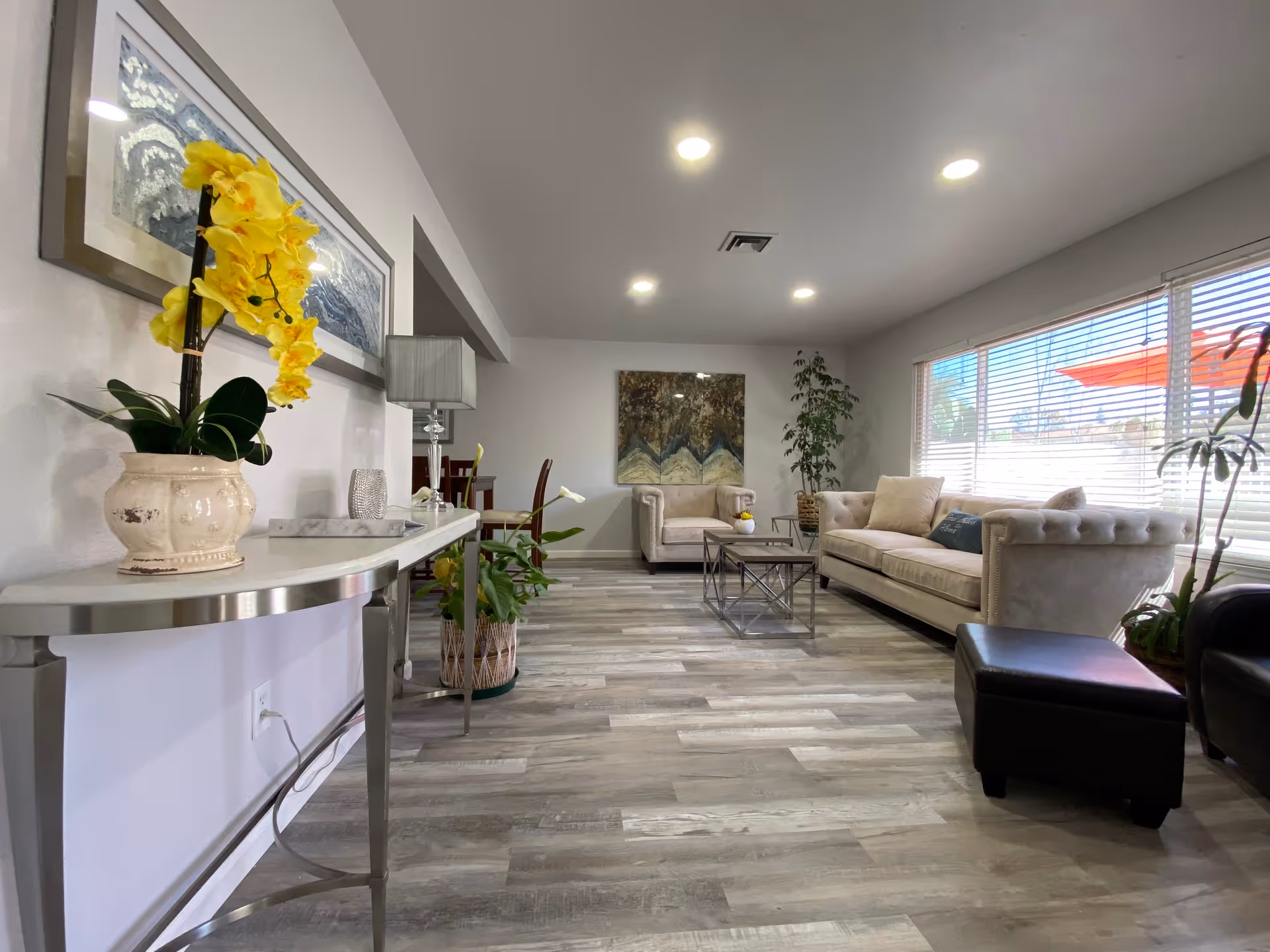 A bright and spacious living room with light gray walls and wood-look flooring. The room features a beige tufted sofa and matching armchair, a glass coffee table with a metal frame, several potted plants, a console table with a yellow orchid, a lamp, and framed artwork on the walls. Large windows with blinds allow natural light to fill the space.