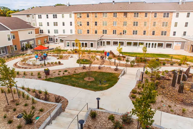 A landscaped outdoor courtyard area of a senior living facility with paved walkways, small trees, shrubs, and seating areas including a table with a red umbrella. The multi-story building surrounds the courtyard with numerous windows and a covered walkway.