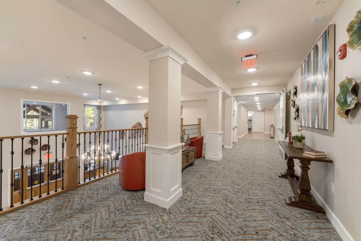 Bright carpeted interior hallway and seating area in a senior living facility with decorative railing, artwork, and a view down to a chandelier-lit entry.