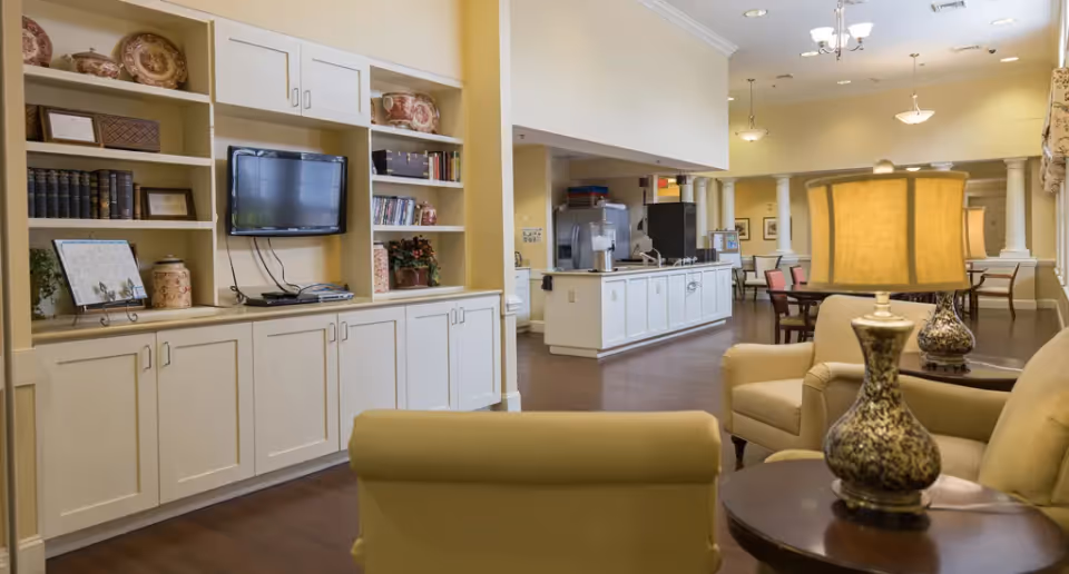 A cozy living room area in a senior living facility featuring beige armchairs, a round wooden side table with a decorative lamp, and built-in white shelves with books, decorative plates, and a mounted flat-screen TV. In the background, there is a kitchenette area with white cabinets and a coffee machine, and a dining area with tables and chairs under warm lighting.