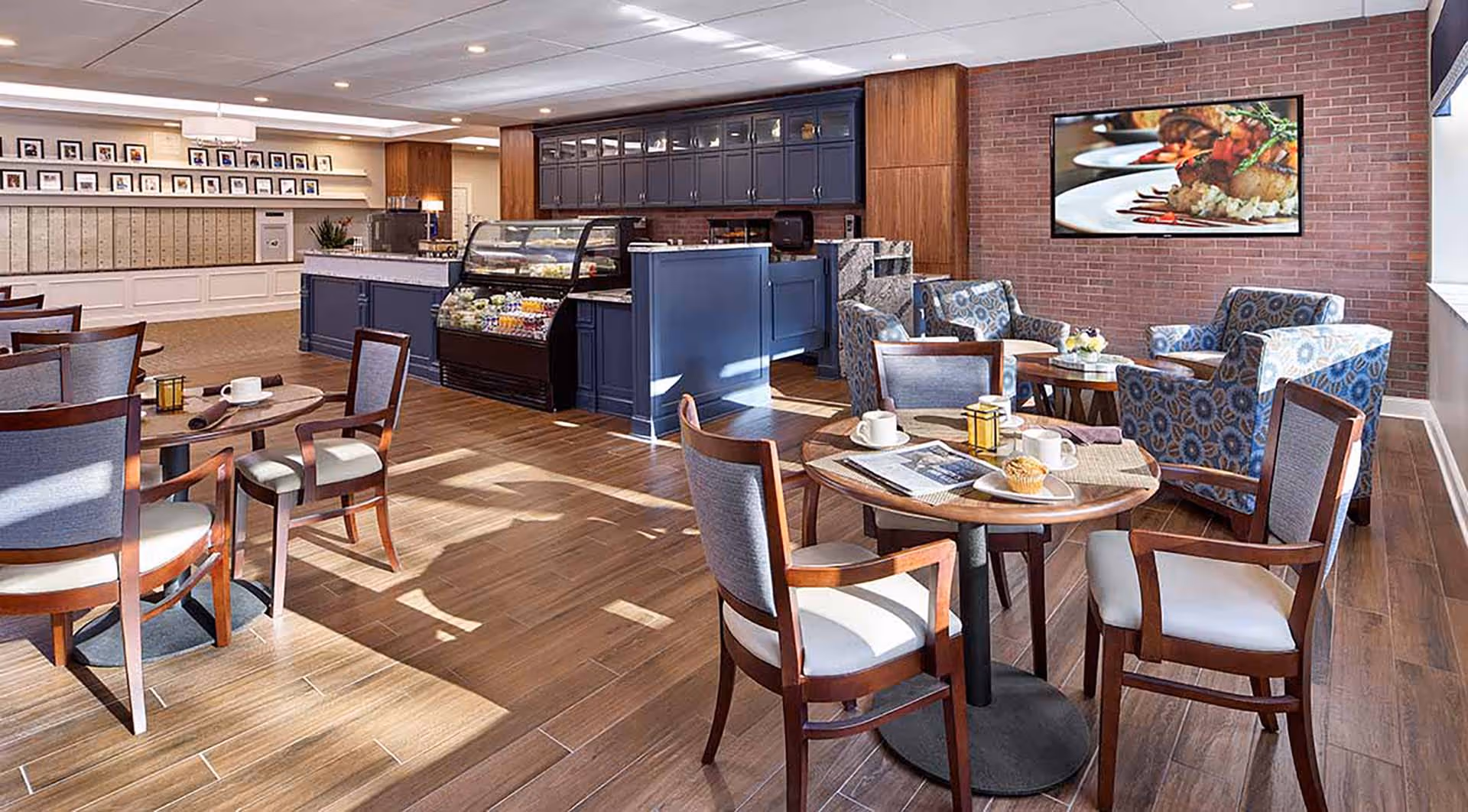 A bright and spacious dining area with wooden floors, several round tables with chairs, and a seating area with patterned armchairs. There is a counter with a display case containing food items, and a large screen on a brick wall showing a close-up image of plated food.