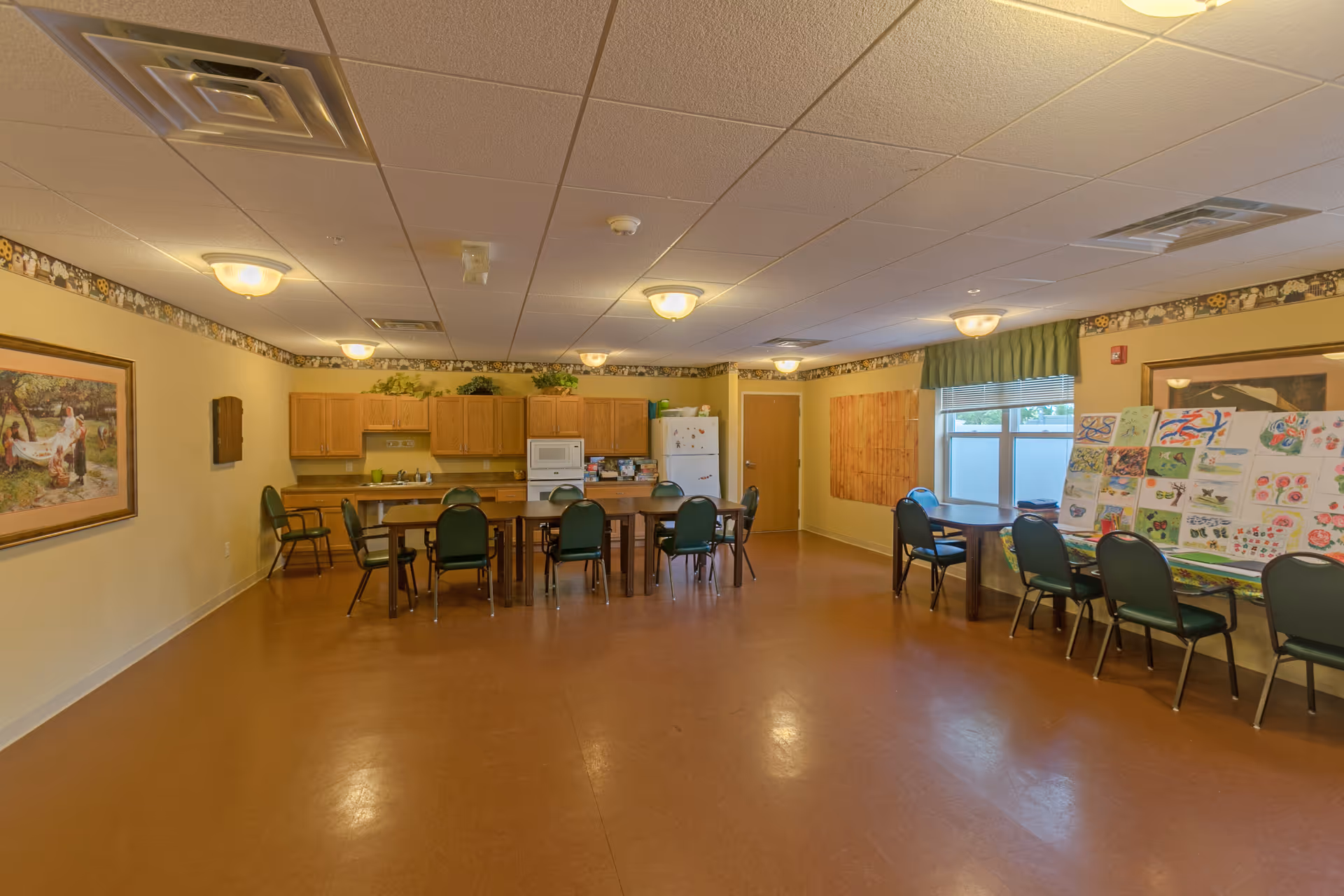 A spacious room with a kitchen area featuring wooden cabinets, a refrigerator, and a microwave. Several green chairs are arranged around tables in the center and along the walls. The room has a brown floor, beige walls with decorative border trim near the ceiling, and multiple ceiling lights. Artwork and colorful drawings are displayed on the walls and tables.