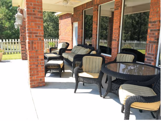 Covered outdoor patio with wicker seating, cushioned chairs and a round glass-top table along a brick house and white picket fence.