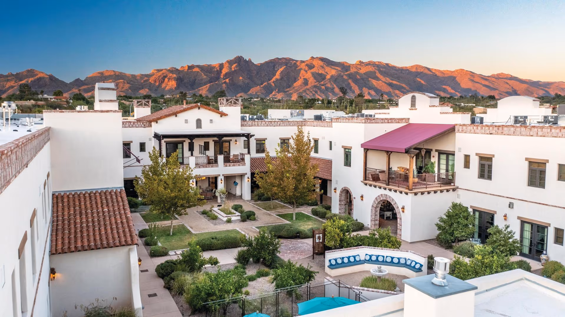 View of a senior living facility courtyard with white buildings featuring red tile roofs and balconies, landscaped greenery, and a seating area with a fountain, set against a backdrop of mountains under a clear sky at sunset.