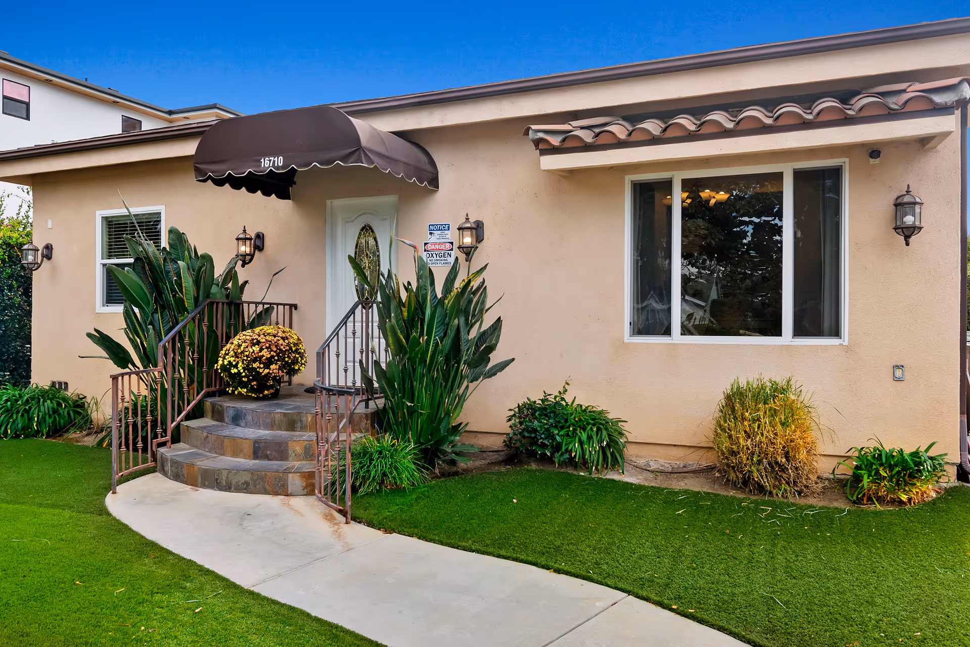 Front entrance of a single-story beige building with a brown awning above the door displaying the number 16710. There are steps leading up to the door with metal railings on both sides, surrounded by green plants and bushes. A concrete pathway curves in front of the entrance, and there is a window to the right of the door with a small overhang above it. Outdoor wall lanterns are mounted on either side of the door and window.